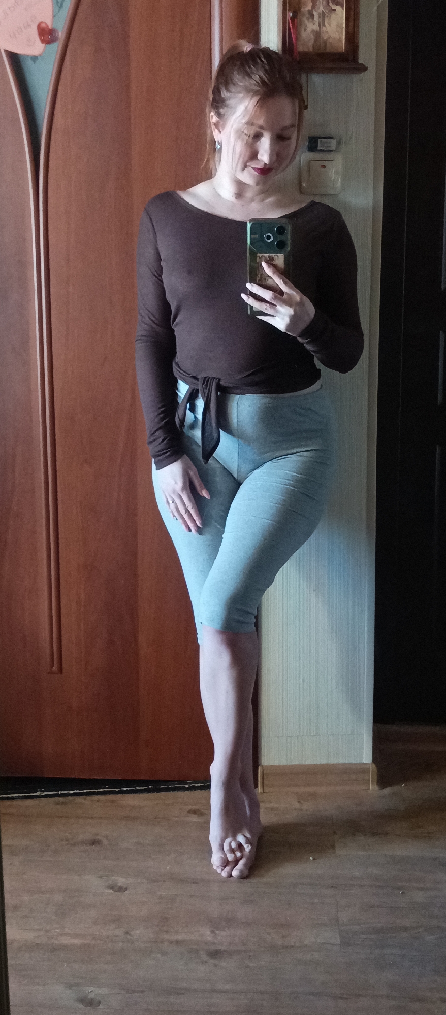 A woman is depicted indoors, taking a mirror selfie. She stands with her left leg crossed over her right, leaning against a light-colored wall on her right side, with her bare feet resting on a wooden floor. Her reddish-brown hair is pulled back, and she is wearing a dark brown long-sleeved top tied at the waist and light grey capri-length leggings. She holds a smartphone with a prominent green case in her right hand, angled to capture her reflection. A ring is visible on her left ring finger, and a small earring adorns her left ear.
To her left, a light brown wooden door with vertical panels is visible, featuring a decorative red heart. Above and behind her right shoulder, a framed picture or icon hangs on the wall, with a wall-mounted object (possibly an electrical outlet or switch) directly below it. A darker vertical structure, possibly another door or piece of furniture, is partially visible on the far right. The setting suggests a home interior, illuminated by what appears to be natural daylight. The woman maintains a calm, neutral expression, looking down towards her phone. No discernible text is clearly visible in the image.