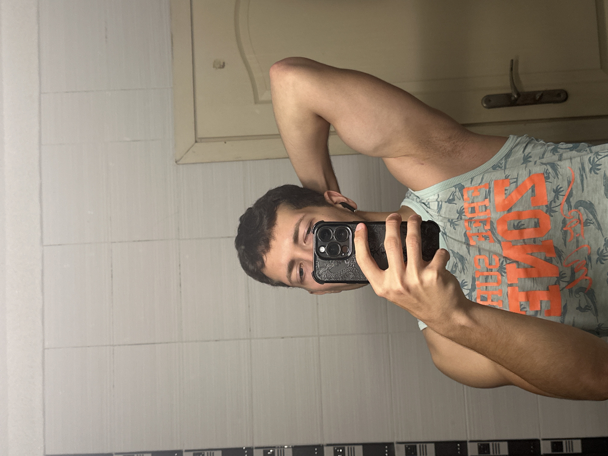 A young man is taking a mirror selfie in a bathroom. He is wearing a light blue tank top with an orange graphic design featuring palm trees and waves. The tank top has the text "THE SOME" visible on it. He has short, dark brown hair and is holding a black smartphone with a patterned case in his right hand, angled to capture his reflection. His left arm is raised, with his hand behind his head, flexing his bicep. He is wearing black wireless earbuds. The bathroom has light-colored tiled walls with subtle horizontal lines. To the right of the man, a portion of a beige door with a dark metal handle is visible. The overall lighting is bright, suggesting the photo was taken during the day or with artificial light.
