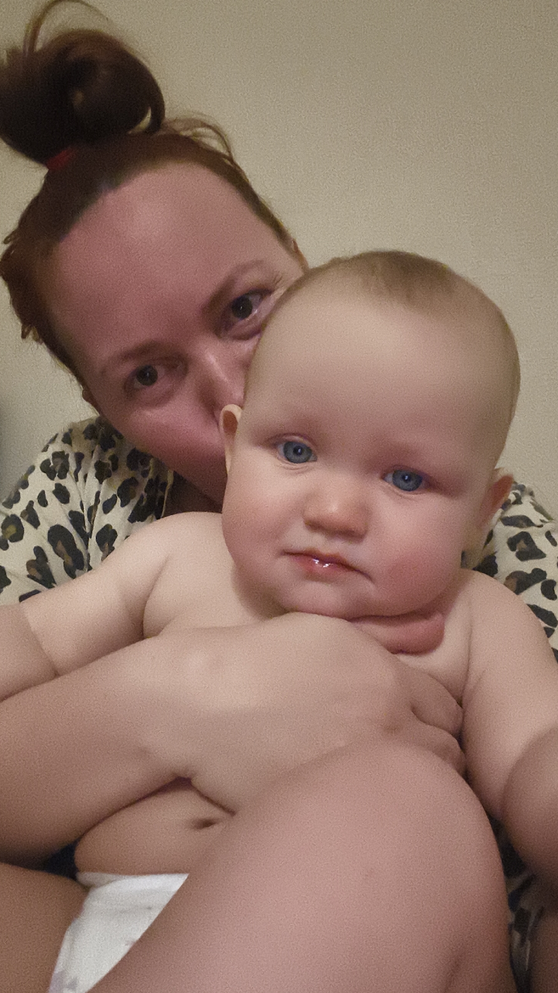 A medium shot depicts a baby with light skin, blue eyes, and a slight furrow in the brow, held close to an adult woman. The baby is positioned in the foreground and appears to be unclothed except for a diaper, with the adult's arms wrapped protectively around it. The woman, visible behind the baby, has auburn hair pulled up in a bun. She is wearing a leopard-print top. The background appears to be a plain, light-colored wall. The location is Unknown, Unknown.