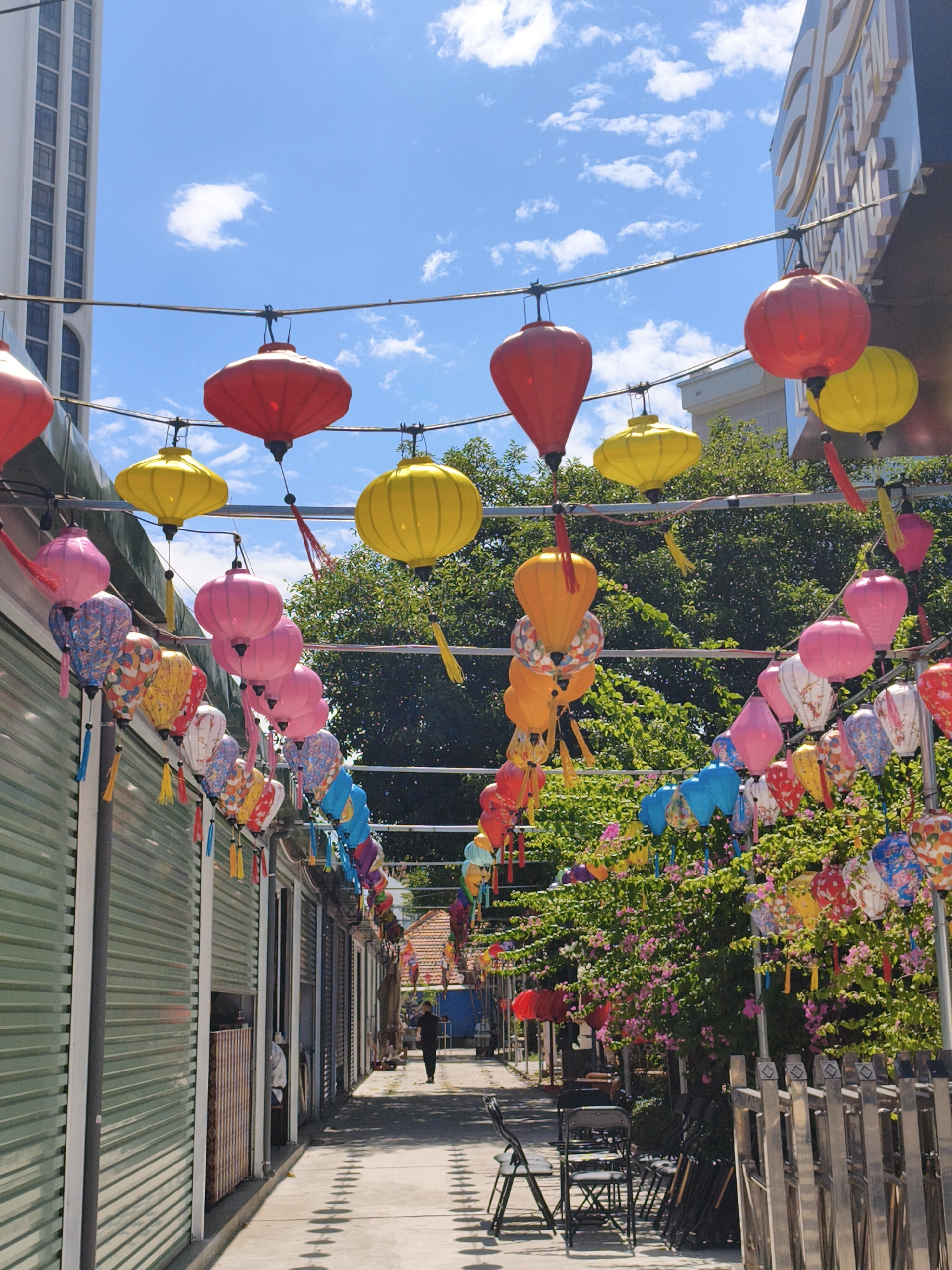 In Nha Trang, Vietnam, a narrow pedestrian alleyway is adorned with a vibrant display of colorful lanterns strung across overhead wires. The lanterns, predominantly red, yellow, and pink, are of various shapes and sizes, some featuring intricate patterns. They hang above a walkway lined with shuttered storefronts on the left and lush green foliage with pink flowers on the right. The ground of the alleyway is paved and shows a repeating pattern of shadows cast by unseen objects. A lone individual, dressed in dark clothing, walks away from the viewer down the alley. In the background, modern buildings and a glimpse of a traditional tiled roof are visible against a bright blue sky with scattered white clouds. The scene suggests a sunny day in a bustling, possibly market, area.