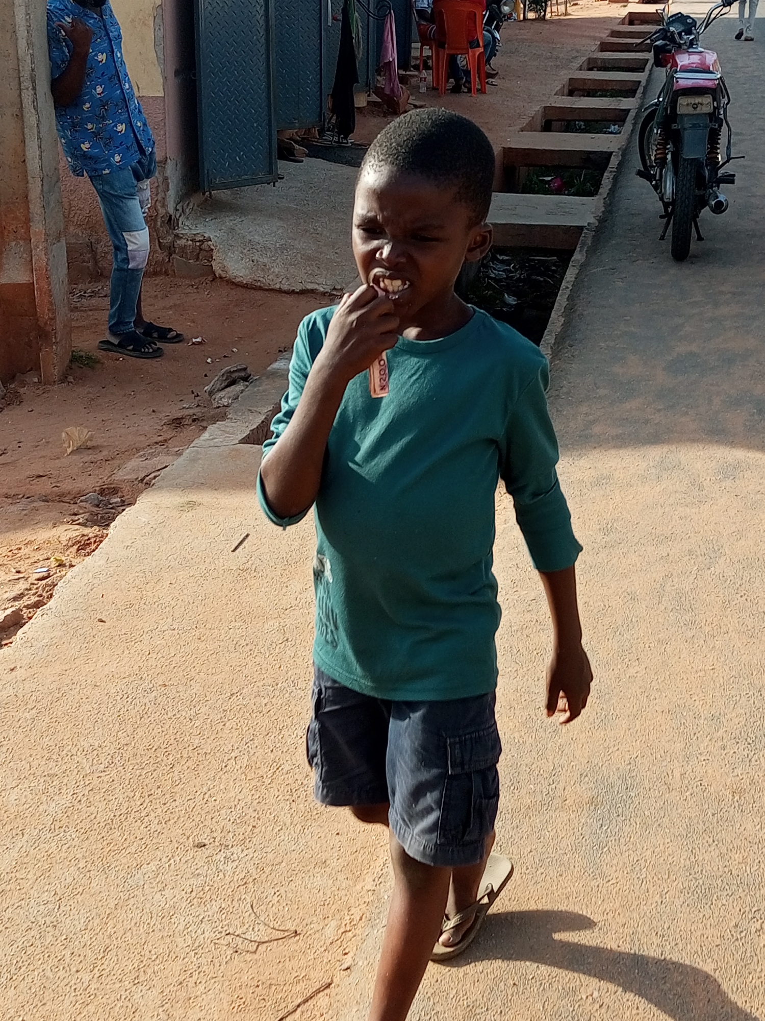 A young boy with short, dark hair walks on a sandy path in Gombe, Nigeria. He is wearing a teal long-sleeved shirt and dark blue cargo shorts. The boy is holding a small, rectangular object to his mouth, appearing to be chewing on it. His expression is focused. To his left, further down the path, an adult wearing a blue patterned shirt and jeans stands near a building with a metal door. Behind the boy, a motorcycle is parked on the paved road, and further in the background, there are orange chairs and other unidentified figures. The environment suggests an outdoor urban setting with a mix of dirt and paved surfaces.