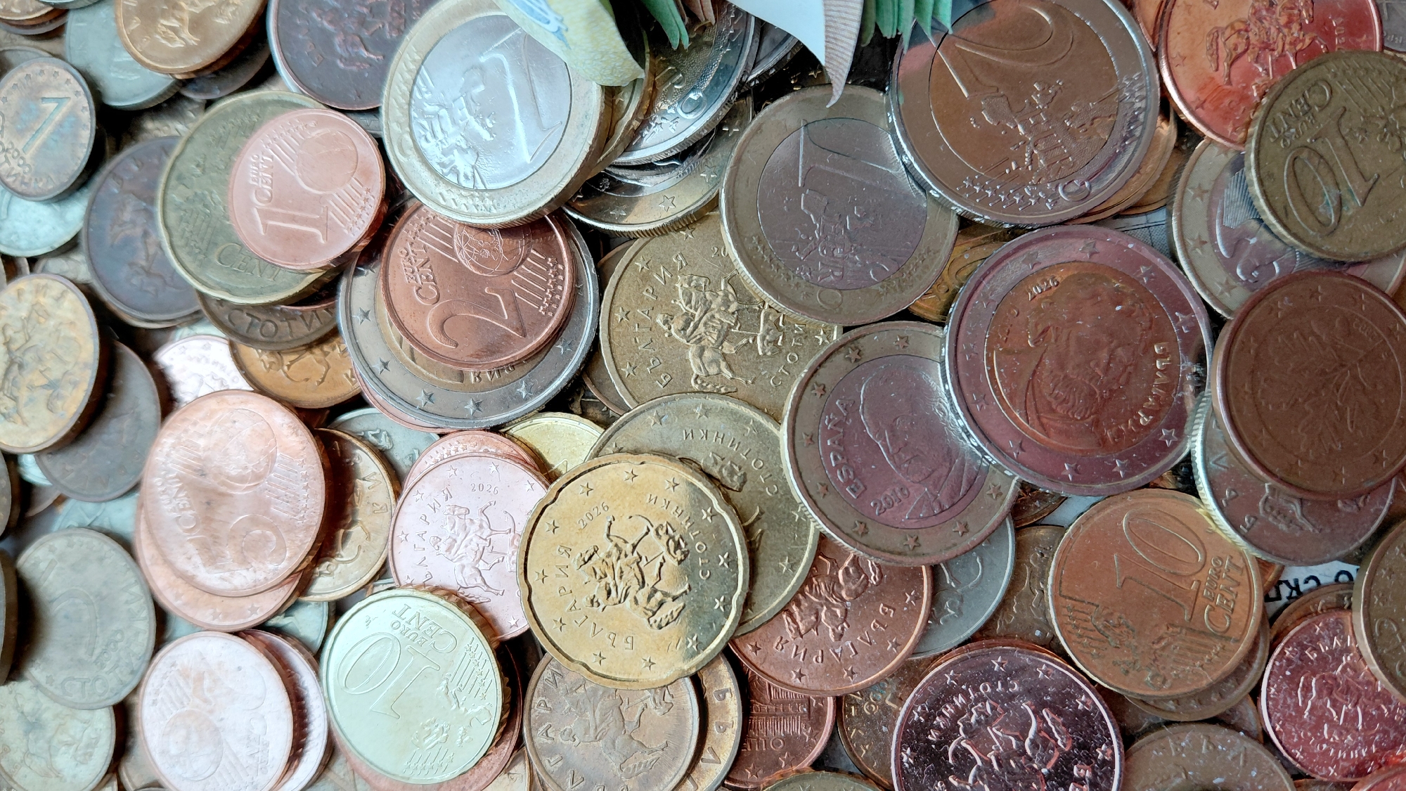 The image shows a close-up of a collection of Euro coins of different denominations. The coins are of varying colors, including bronze, copper, gold, and silver. Some coins have dates printed on them, such as 2016 and 2026. A few of the coins have the word "Bulgaria" printed on them in Cyrillic, and one coin has the word "España" printed on it. Some coins also have "Euro Cent" printed on them.