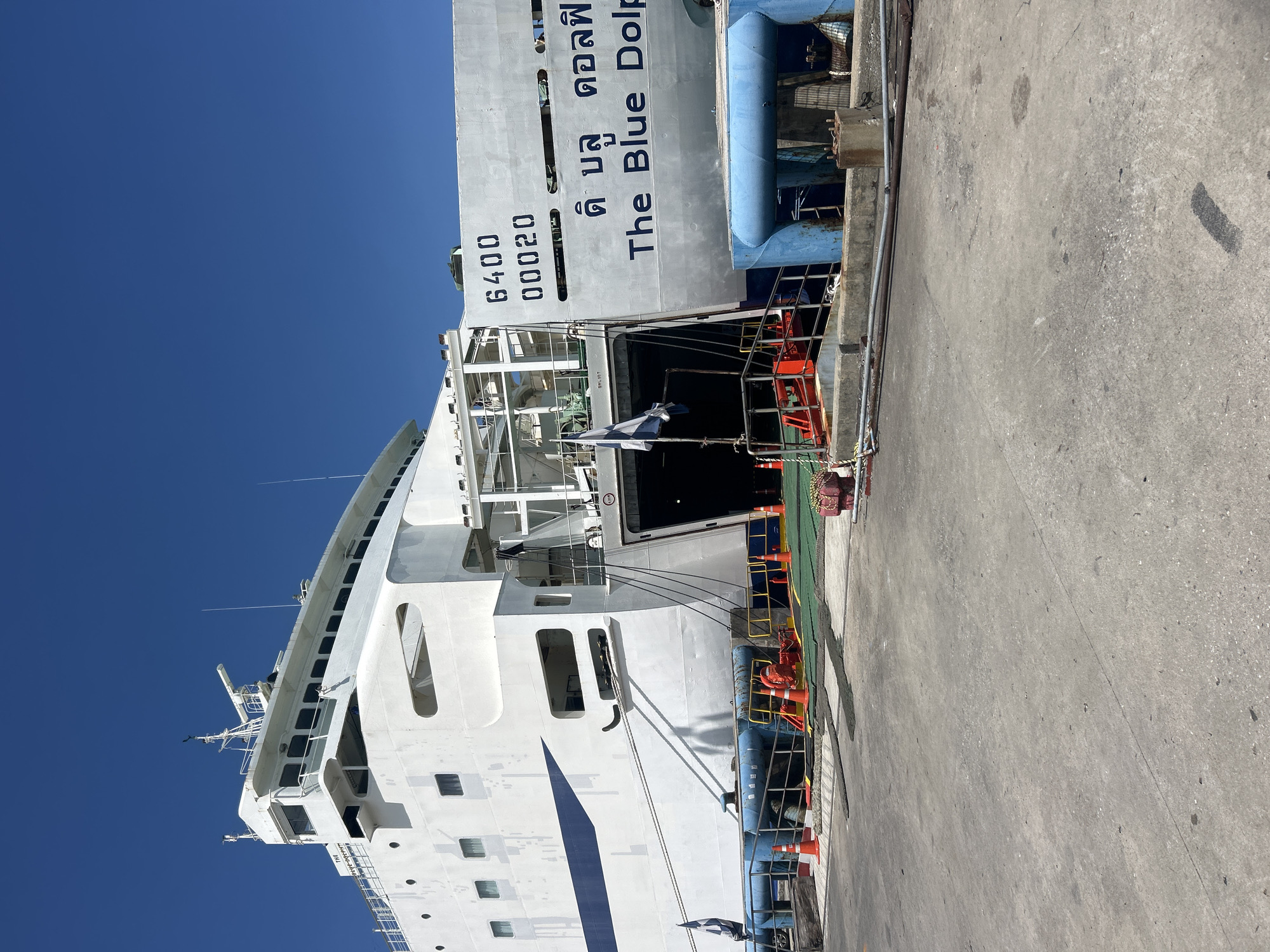 An outdoor, eye-level, full-shot image captures a large white passenger ferry, named "The Blue Dolphin," docked at a port in Ko Samui District, Thailand. The ferry, predominantly white with a blue stripe along its hull, is positioned at a concrete pier under a clear blue sky. Its bow is angled towards the viewer's left, revealing a large, open vehicle ramp at the stern, indicating it is prepared for loading or unloading. The ramp is green on its surface and bordered by a yellow railing. Orange traffic cones are strategically placed on the ramp and the pier, suggesting traffic management.
On the stern of the ferry, large black numbers "6400" and "00020" are visible above the name "The Blue Dolphin," written in both English and Thai script. The Thai script above "The Blue Dolphin" appears to read "ด ี บลู ดอลฟิน". A black and white flag is attached to a pole on the side of the ferry. Several mooring lines are visible, connecting the ship to the pier. Blue bollards, used for securing ships, are situated along the dockside. In the background, the upper decks of the ferry rise towards the sky, showcasing multiple windows and nautical equipment. The overall scene is one of active port operations, with no people clearly visible in the frame.