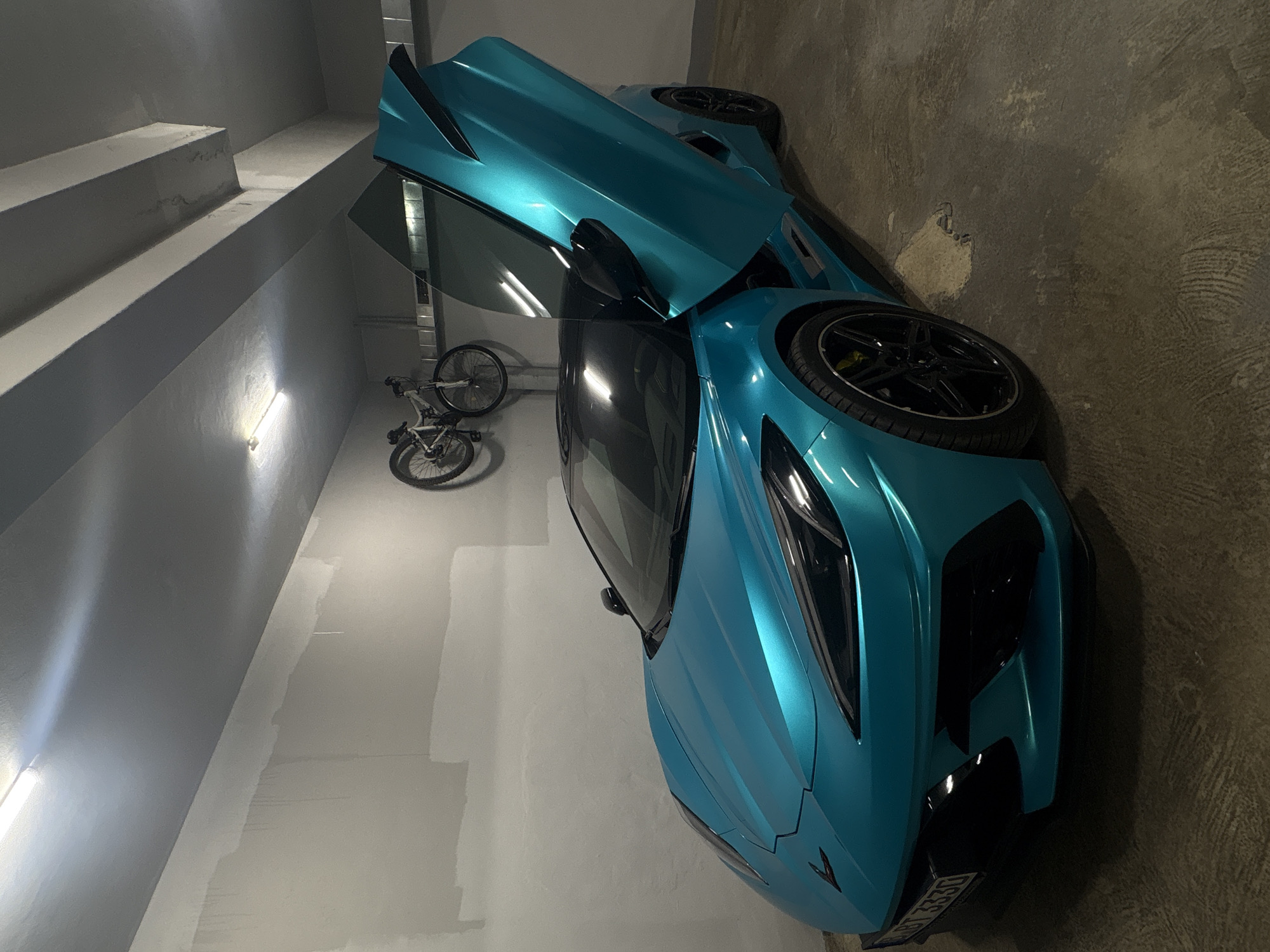 The image shows a turquoise sports car parked in what appears to be a garage or indoor parking area. The car's door is open, revealing part of the interior. A bicycle is leaning against the wall in the background. The walls are painted in shades of gray. There are steps visible to the left. The location is potentially Varna, Bulgaria, although the photo alone provides no definitive proof of this. The lighting is from artificial sources.