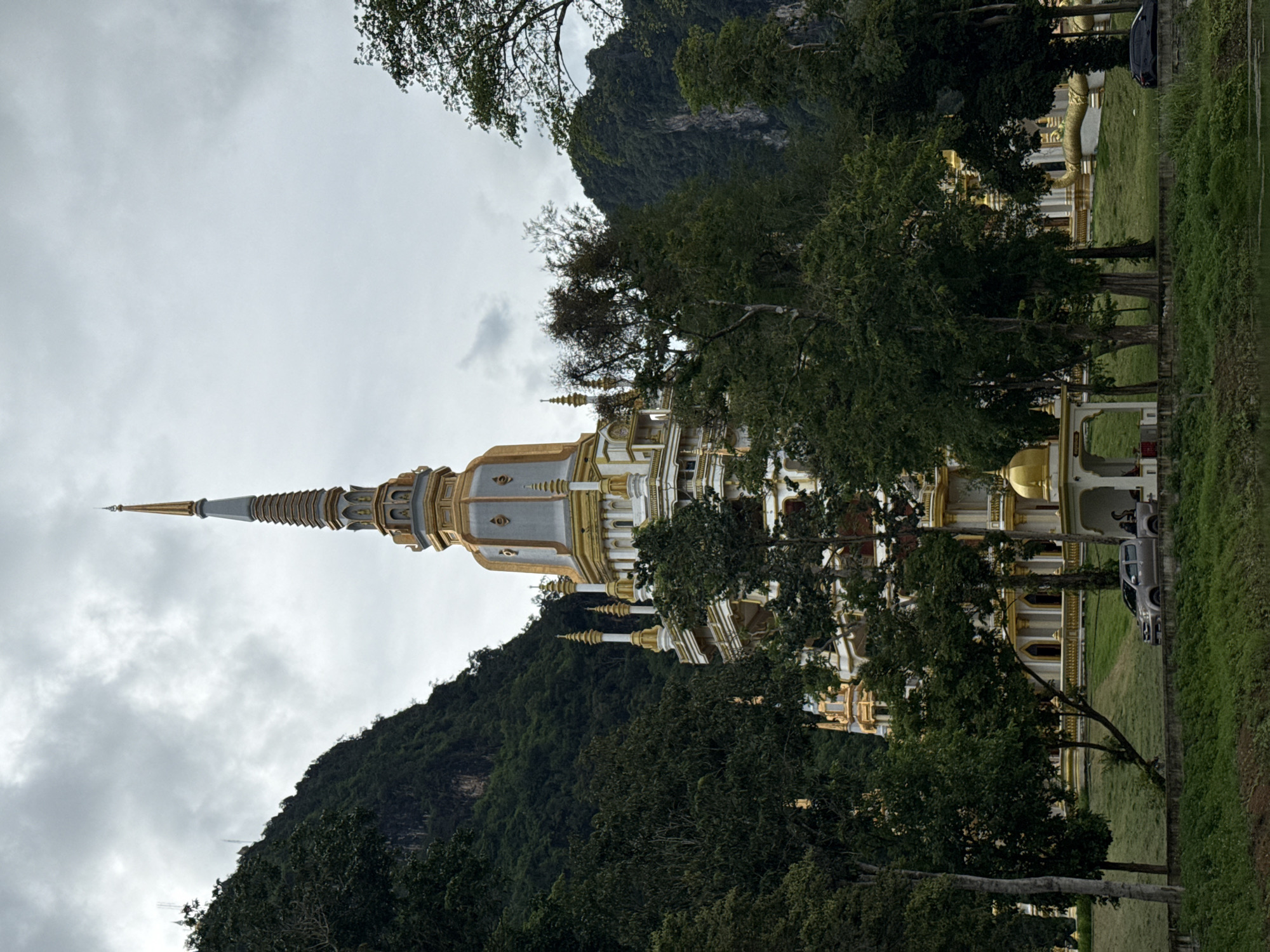 A multi-tiered white and gold stupa stands prominently against a backdrop of lush green mountains and a cloudy sky. The stupa, with its ornate spires and gold accents, is partially obscured by a dense canopy of trees. In the foreground, a body of water reflects the overcast sky. A grey pickup truck is parked on a grassy area in front of the stupa, and a black car is visible further down the road. The scene appears to be in a natural, somewhat humid environment, likely in the region of Ban Khao Tang, Thailand, given the architecture and surrounding landscape.