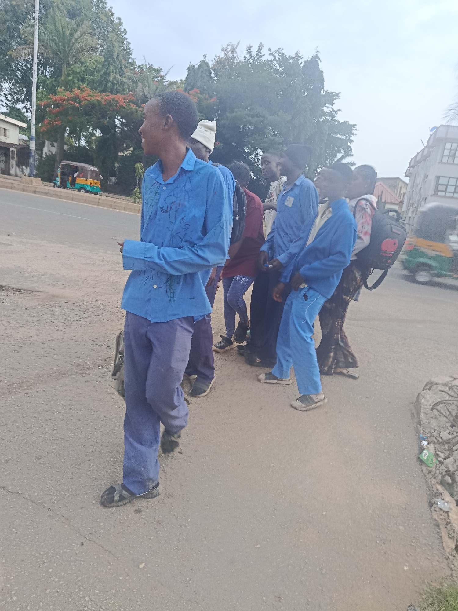 The image depicts a group of young people, likely students, walking along a paved road in what appears to be a semi-urban area. The foreground is dominated by the legs and lower bodies of several individuals, mostly wearing blue shirts and pants. The person closest to the viewer is wearing blue pants and a blue collared shirt with dark markings. Another person behind them wears blue pants and a lighter blue collared shirt. Further back, a person in a red shirt is visible. To the right, a person in a patterned skirt and a blue jacket carries a black backpack.
The background reveals a street with a vehicle, possibly a tuk-tuk, in motion on the left side. Trees line the side of the road, with some bearing red flowers. Buildings are visible in the distance, suggesting a populated area. The sky is overcast. The scene conveys a sense of everyday movement and activity. The location is identified as New Stadium, Nigeria.