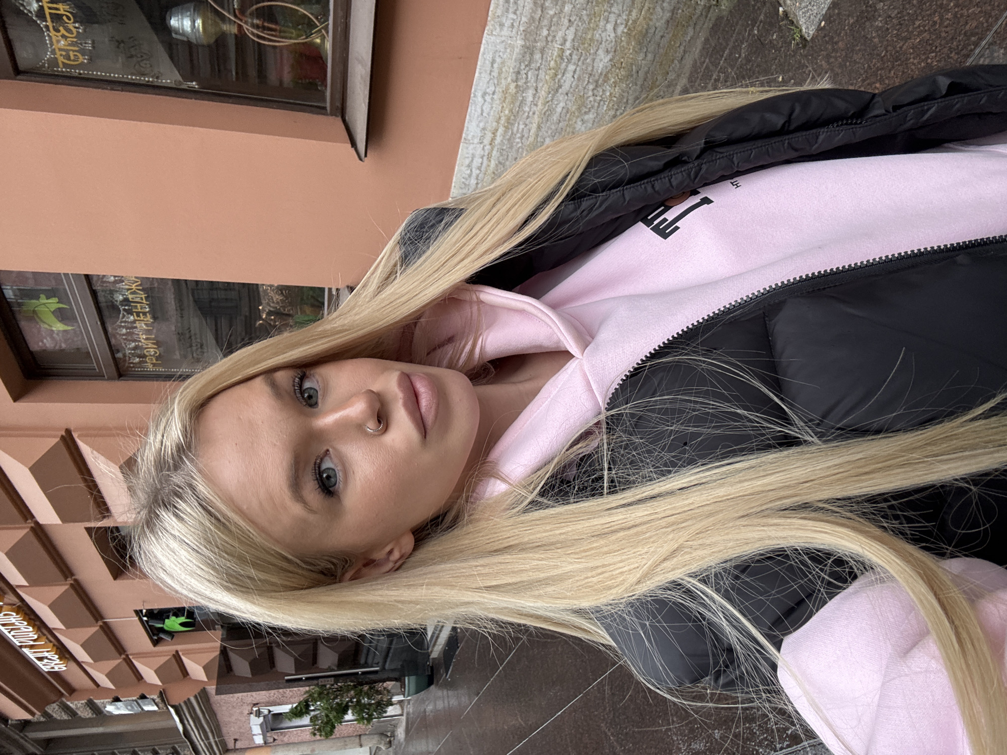 A young woman with long, blonde hair and blue eyes is pictured from the chest up, looking towards the camera. She is wearing a pink hoodie with a black jacket over it. A nose ring is visible on her right nostril. The background shows a building with a salmon-colored facade and a window with lettering that appears to be in Cyrillic script, suggesting a location in Russia, possibly Saint Petersburg given the context. The ground is wet and reflective, indicating it might be raining or has recently rained, consistent with a cloudy or damp day. The overall mood is casual and personal, captured in an outdoor urban environment.