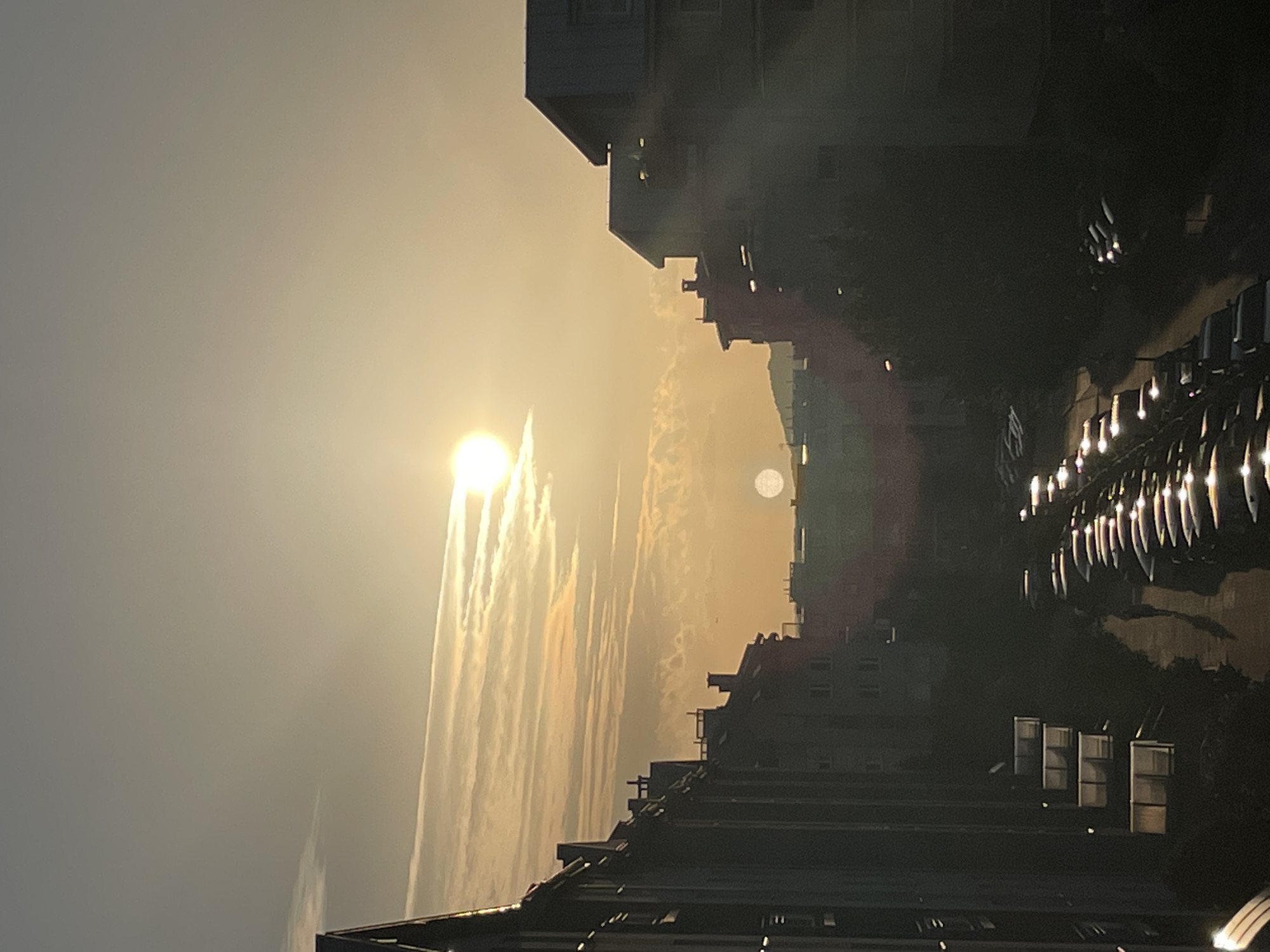 The image depicts a sunset or sunrise over a city. The sun, a bright orb with a luminous halo, is partially obscured by wispy clouds that appear golden and streaky. The sky is a gradient of warm tones, transitioning from a bright yellow near the sun to a muted gray. In the foreground, silhouetted buildings rise against the bright sky, their architectural details softened by the low light. Some buildings have visible windows, and decorative lights are strung along what appears to be a balcony or rooftop, casting a warm glow.
The scene is oriented vertically, with the sun and sky occupying the left side and the city landscape on the right. The buildings are dark silhouettes, suggesting they are either far away or the lighting conditions are such that their colors are not discernible. The presence of multiple buildings and what might be a street lined with parked cars indicates an urban environment.
The overall mood is serene and atmospheric, enhanced by the warm light and the quiet, silhouetted cityscape. The time of day is clearly either dawn or dusk, given the position and appearance of the sun and the quality of light. There is no visible text in the image.