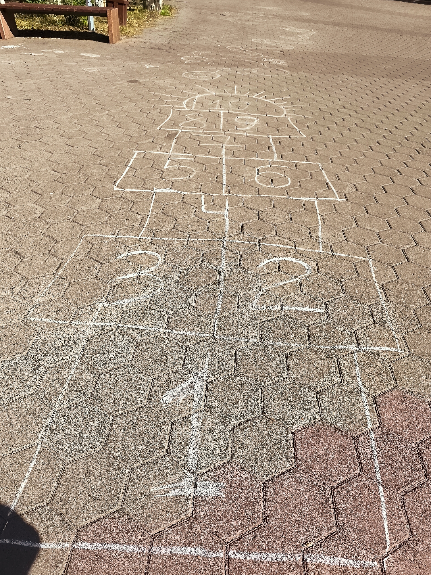 The image shows a section of a paved area, likely a public walkway or playground, in Qarağandı city administration, Kazakhstan. The paving stones are hexagonal and a mix of tan and reddish-brown colors. White chalk lines form a hopscotch grid drawn on the pavement. The grid consists of several squares and rectangles, some of which are numbered. In the foreground, there are squares numbered "1", "2", and "3". Further up the grid, there are more squares with indistinct markings. To the left and in the background, a wooden bench with metal supports is visible. The area is outdoors and appears to be a sunny day, casting shadows on the pavement. The scene depicts a recreational area prepared for a game of hopscotch.