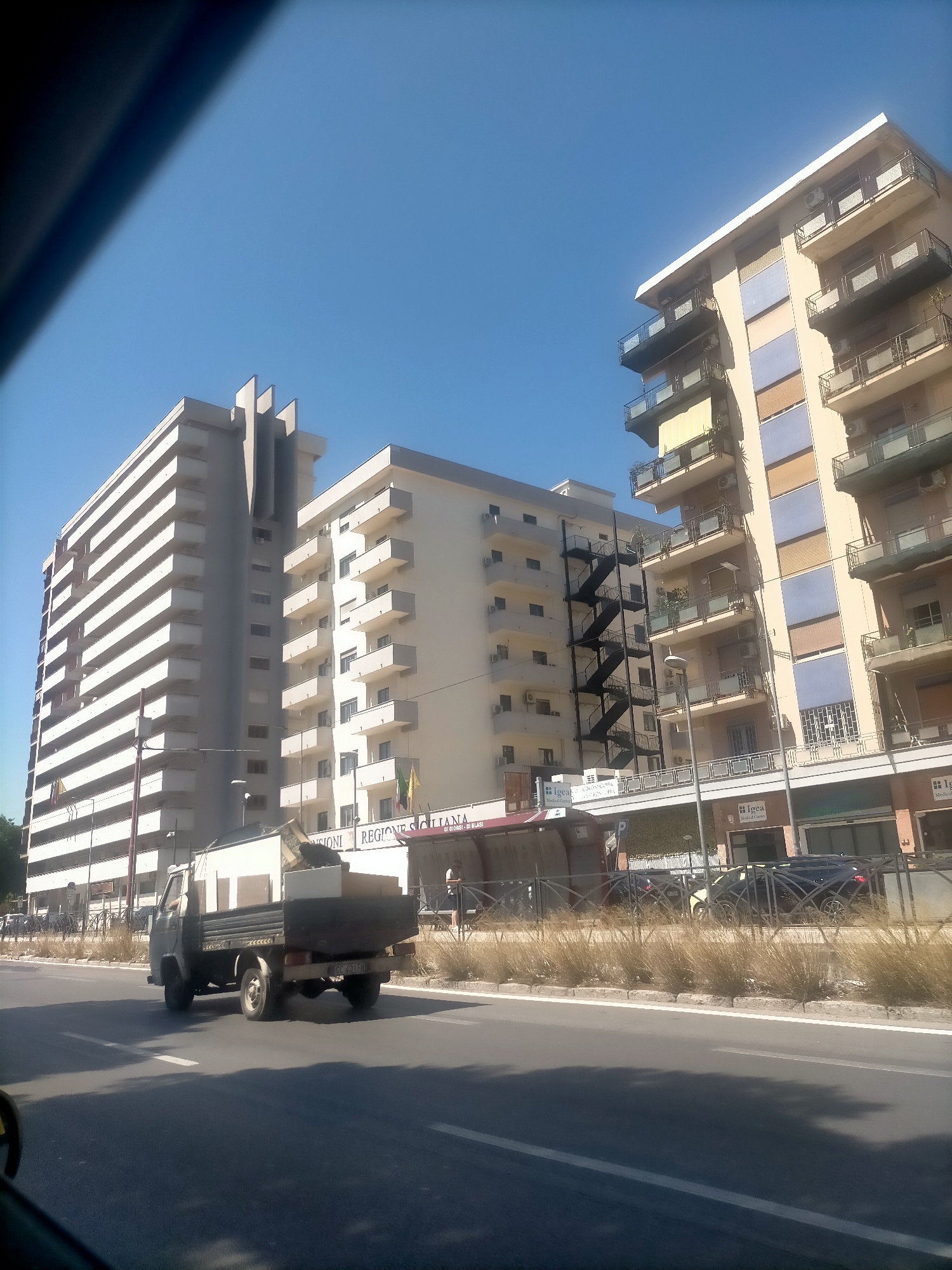 The image shows a street scene in Palermo, Italy. In the foreground, a truck is driving on the road, loaded with furniture. The truck is light gray with a darker gray bed. Behind the truck, there are modern apartment buildings. One building is tall and white with many balconies. Another building is shorter and has a cream-colored facade with balconies and some colorful panels. A metal fire escape is visible on the side of one of the buildings. The sky is clear and blue. There are some businesses visible on the ground floor of the buildings, including one with a sign that says "Igea Medical Center". The overall impression is an urban environment on a sunny day.