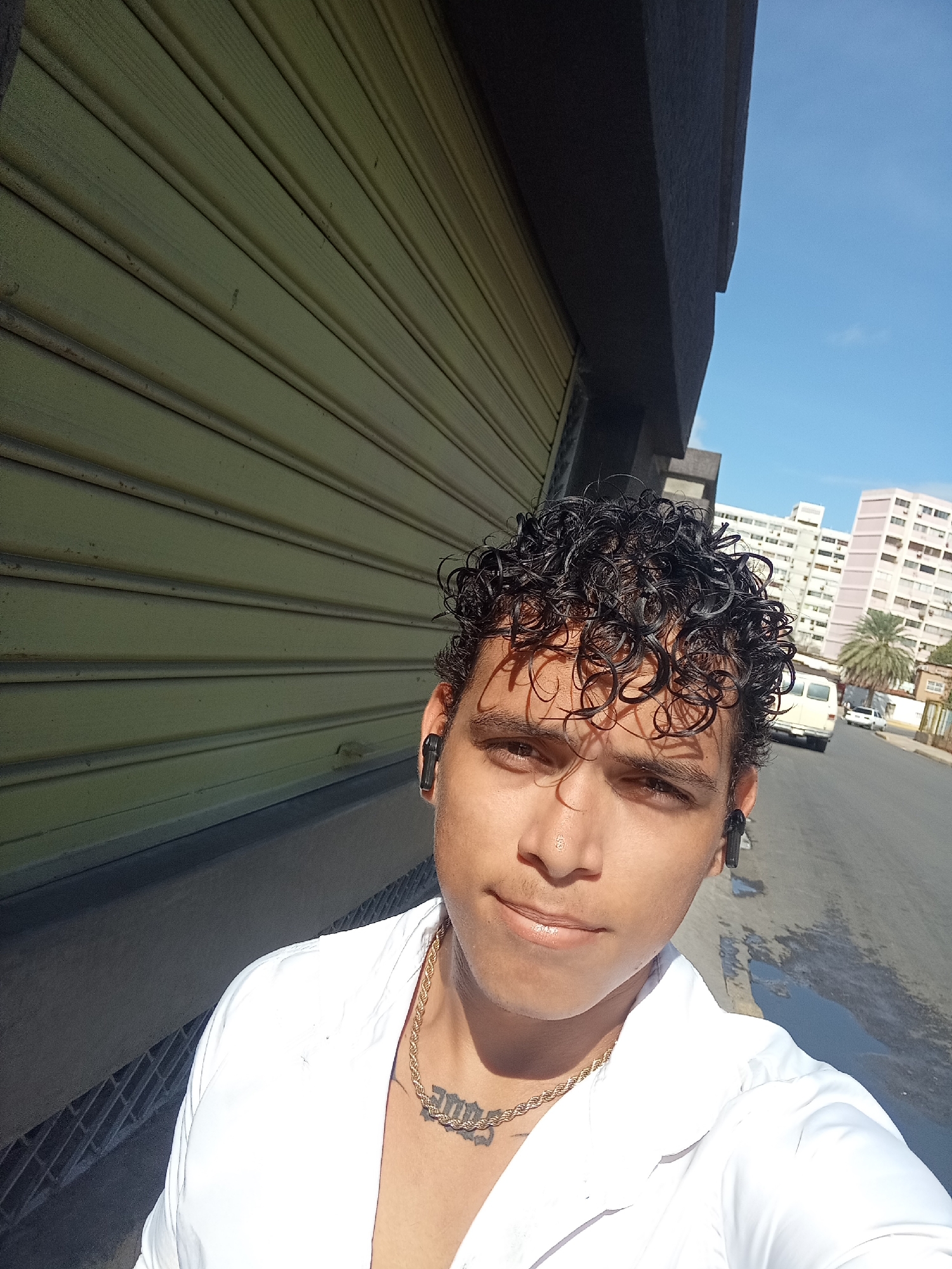 The image is a close-up, self-taken photograph of a young man with curly dark hair and brown eyes. He is wearing a white collared shirt and a gold chain necklace with a pendant that appears to be an inscription or symbol. He has black wireless earbuds in both ears. The man is standing outside in what appears to be an urban environment, possibly Juan Griego, Venezuela. Behind him, a building with a large, closed, light yellow corrugated metal shutter dominates the left side of the frame. The right side of the image shows a street with vehicles, including a white van and a white car, and tall buildings in the background under a clear blue sky. The ground appears wet in some areas, suggesting recent rain. The man is looking directly at the camera with a slight smile.