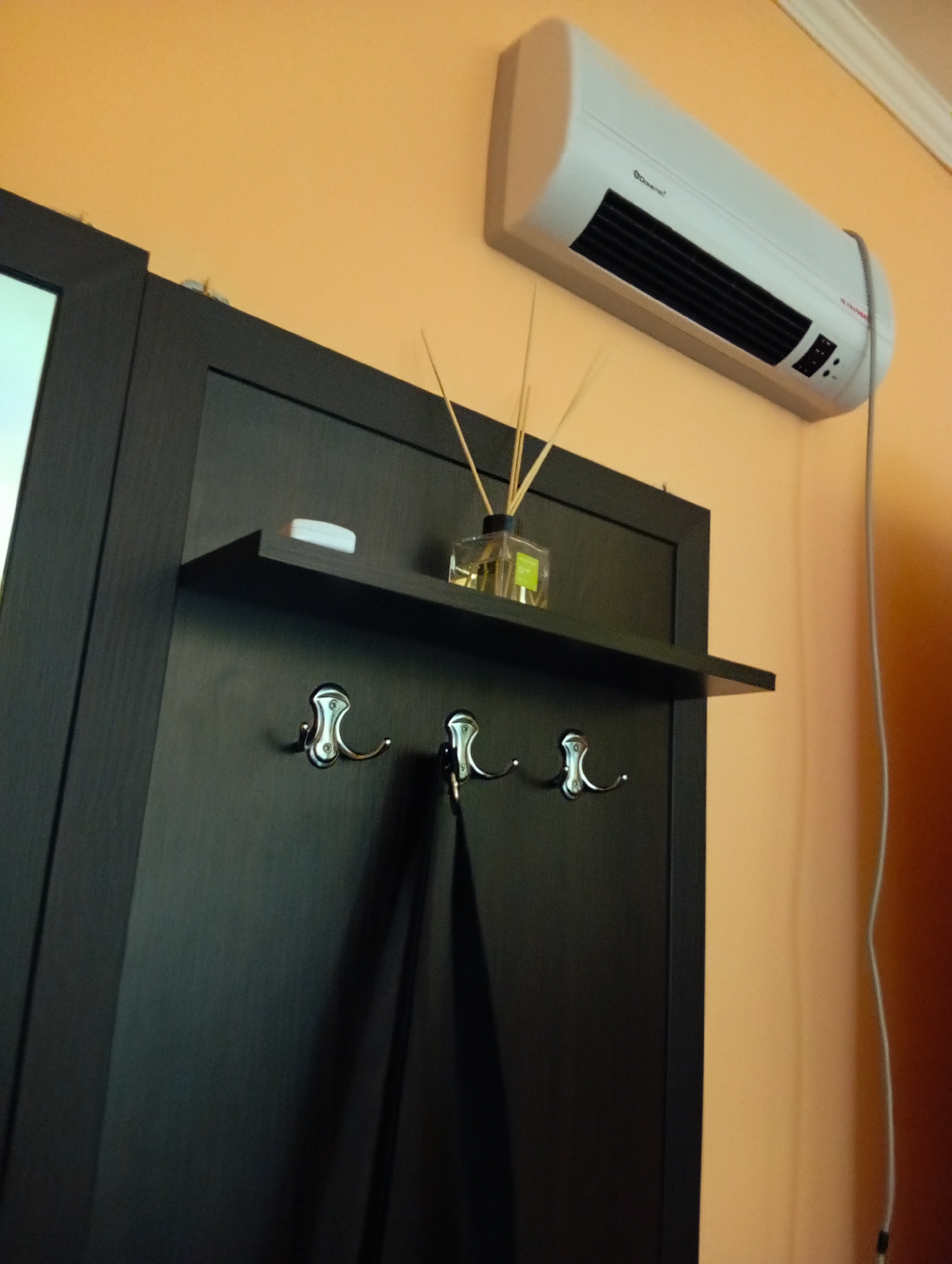 The image shows a segment of an interior wall in Poltava, Ukraine. The wall is painted a light orange color. Mounted high on the wall is a white, cylindrical-shaped electric heater. Below the heater and offset to the left is a dark-colored coat rack or hallway organizer. The coat rack has a small shelf with a clear glass bottle containing reed diffusers, as well as a small white object, possibly a bar of soap. Three silver-colored hooks are mounted below the shelf. A dark-colored garment, perhaps a coat or jacket, hangs from the middle hook. The left edge of the coat rack adjoins what appears to be the frame of a mirror. A white power cable descends from the heater along the wall.