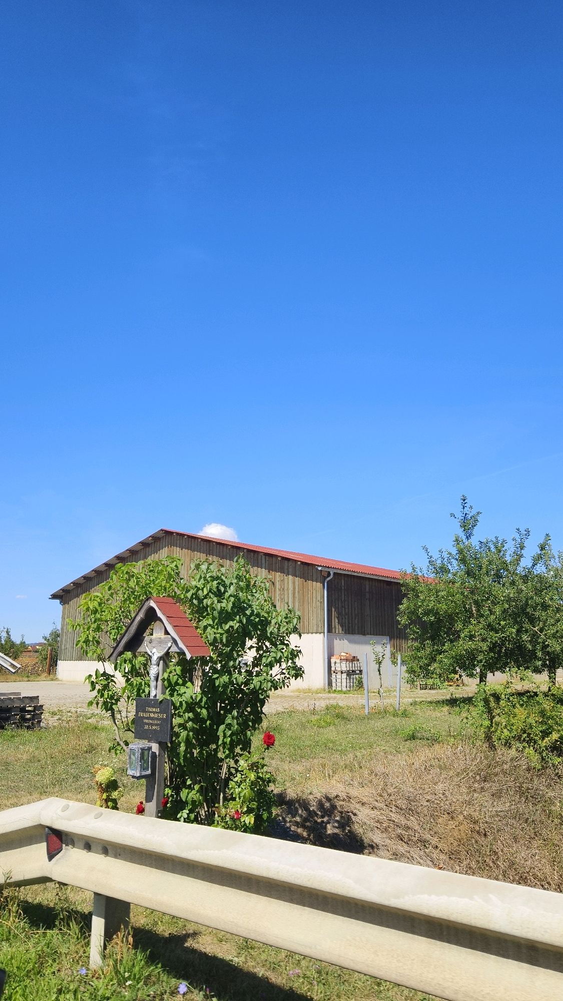 The image depicts a roadside shrine next to a large wooden barn under a clear blue sky in Feuersbrunn, Austria. The shrine, adorned with red roses, features a crucifix with a figure of Jesus Christ and a plaque with the name "THOMAS FRAUENWIESER" and dates "GEB. 28.9.1985 VERSTORBEN 20.9.2015". A small lantern is also present at the base of the shrine. The barn, constructed of vertical wooden planks with a reddish roof, is situated behind the shrine and appears to be a utilitarian structure. The surrounding environment consists of dry grass, a few green trees, and a concrete guardrail in the foreground. A hint of another building can be seen to the left, and stacks of bricks or building materials are visible against the barn. The scene is bathed in bright sunlight, casting shadows on the ground.