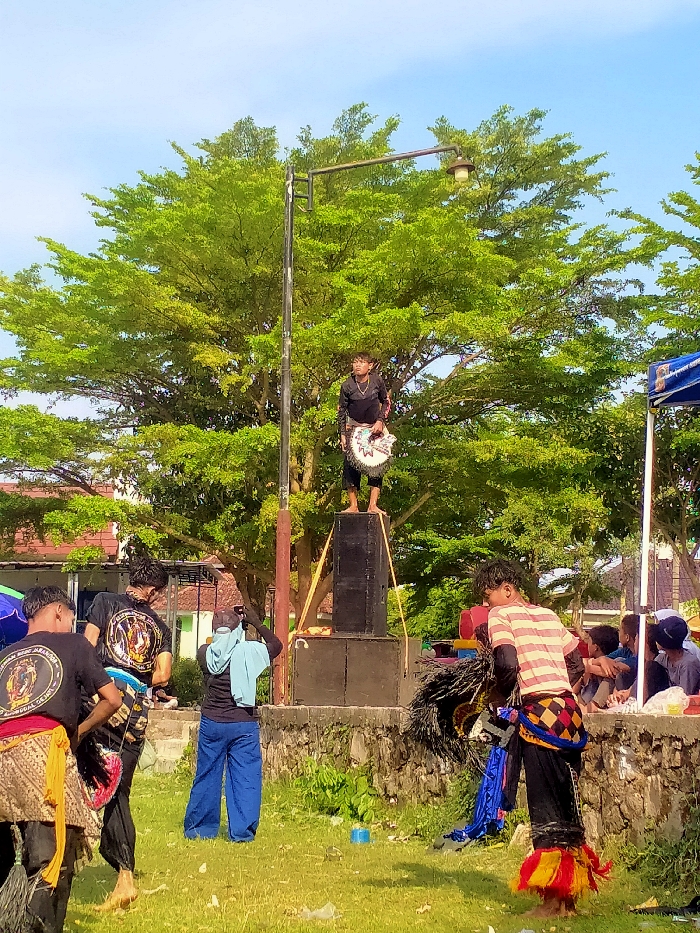 The image depicts an outdoor cultural performance or event in the city of Jetak, Indonesia, under a clear blue sky. A young male, barefoot and dressed in a black long-sleeved top and dark trousers, stands balanced on two vertically stacked black speakers, holding a white and black fan-like prop with a decorative edge. This central figure is positioned directly beneath a tall metal streetlamp pole.
In the immediate foreground, to the left, two males are visible from behind, wearing black t-shirts adorned with yellow and white circular emblems. One of them wears a yellow/brown sarong-like garment over black trousers. Further back, another individual, seen from behind, wears a black shirt, blue trousers, and a light blue cloth draped over their head and shoulders, appearing to photograph or record the event with a device held at eye level. To the right, another male in a red-and-white striped t-shirt, black trousers, and elaborate traditional blue, yellow, and red leg and waist coverings stands, holding a dark, feathered or furry object. On the far right, several individuals are partially visible, seated under a blue canopy tent.
The ground is a grassy field with some visible litter. A low stone wall separates the performance area from a slightly elevated section in the midground. Large, lush green trees form a dense background, with residential buildings featuring red-tiled roofs faintly visible beyond them. The scene is brightly illuminated by natural sunlight, casting visible shadows.