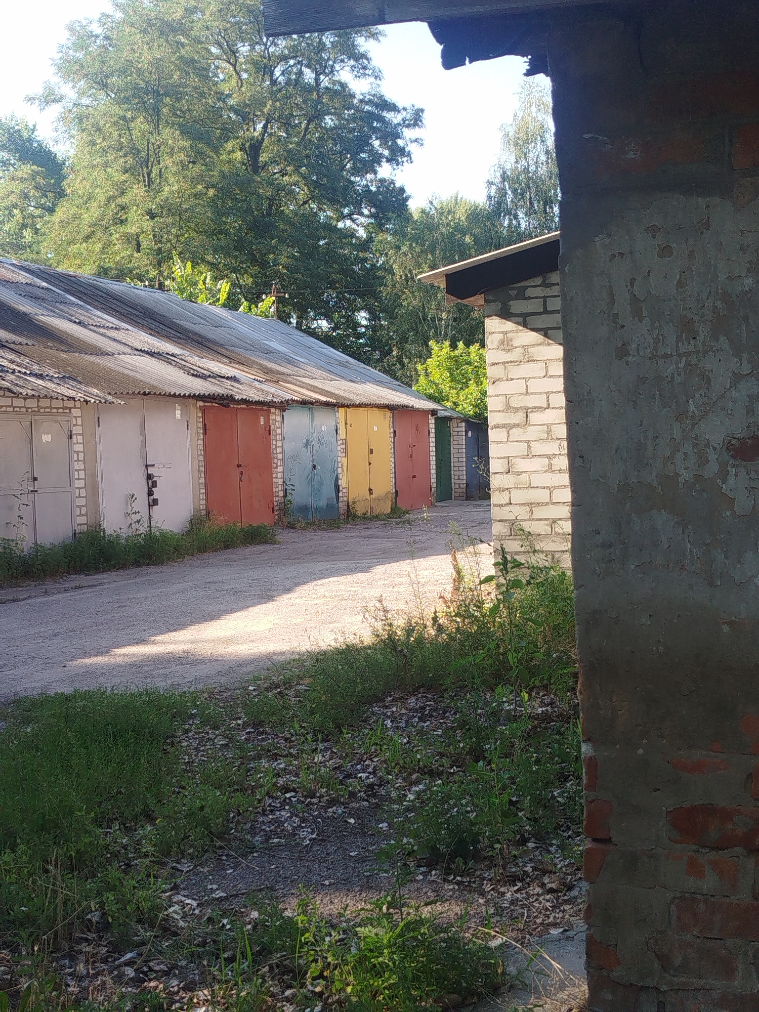 This is a photograph taken in Kremenchuk, Ukraine. The image depicts a row of garages with corrugated metal roofs, set against a backdrop of trees and a clear sky. The garages are of varying colors, including gray, red, blue, and yellow. In the foreground, a gravel path leads towards the garages, with overgrown green grass and foliage on either side. A brick structure with peeling paint is visible on the right edge of the frame, partially obscuring the view of the garages. The lighting suggests it is daytime, with sunlight casting shadows on the path. There are no people or animals visible in the image. The scene appears to be an outdoor storage area or parking lot in a residential or industrial district.