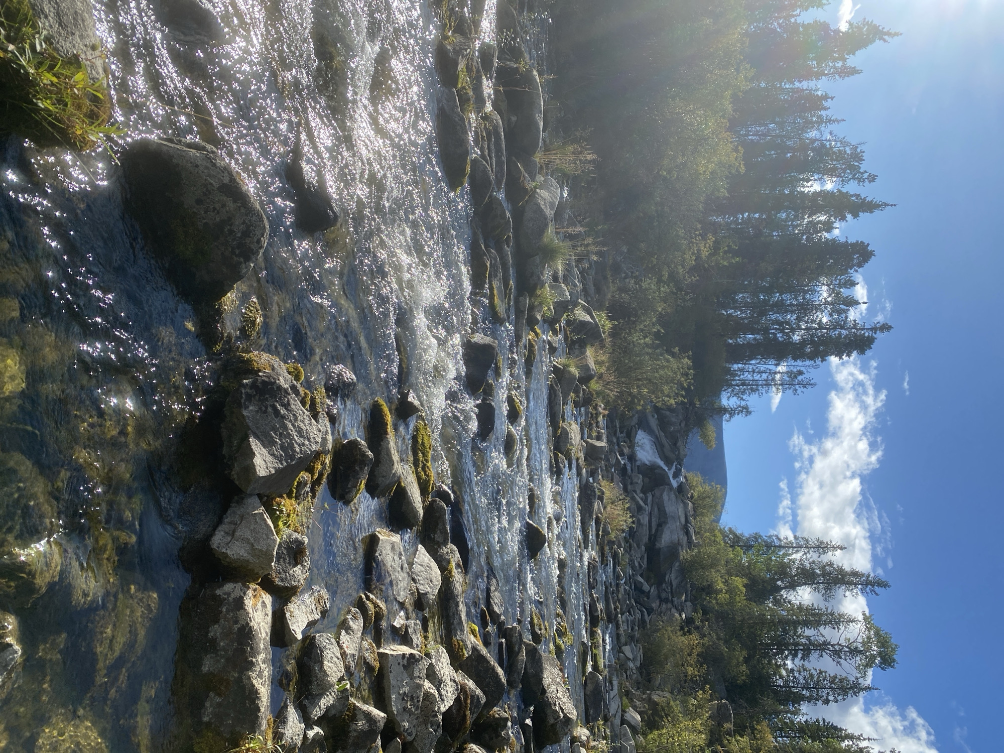 The image depicts a sun-drenched, rocky riverbed in a mountainous region. Tall, dark green conifer trees line the upper slopes of the scene, interspersed with patches of lighter green foliage. The sky is a vibrant blue, dotted with fluffy white clouds, and a bright sun casts a strong lens flare from the upper left corner. A clear, shallow river flows over numerous rounded, grey rocks, many of which are partially submerged. The water is turbulent in some areas, creating ripples and reflecting the sunlight. In the background, a small waterfall cascades down a rocky incline. The overall impression is of a natural, undeveloped landscape. The location is specified as Yukon, Unorganized, Canada.