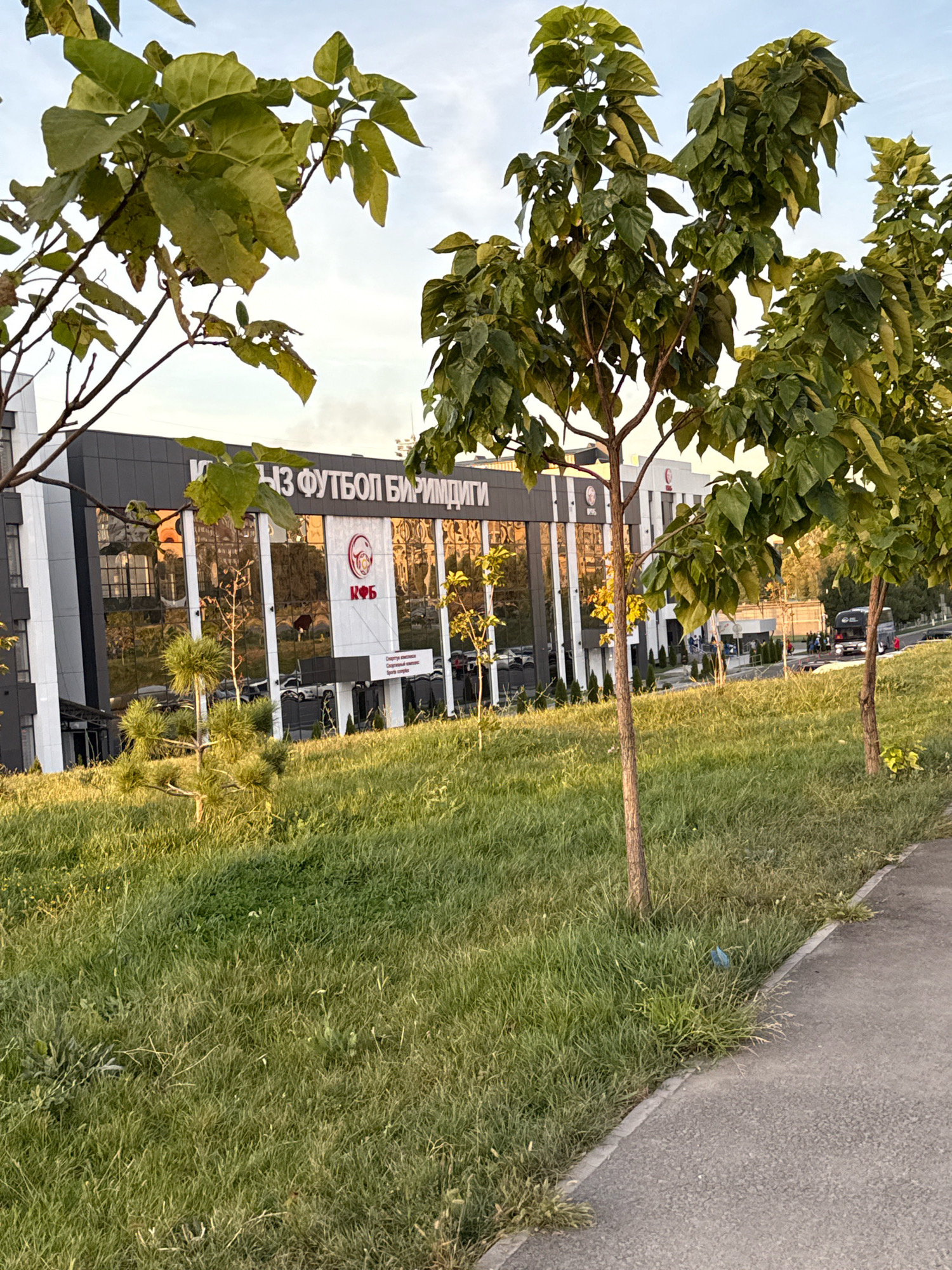 In Bishkek City, Kyrgyzstan, a building with a dark facade and mirrored windows, identified by Cyrillic lettering that reads "ФУТБОЛ БИРИМДИГИ" and a logo with the abbreviation "КФБ", stands behind a grassy slope. The building appears to be a sports facility, possibly a football club. A bus is parked in front of the building, and a group of people are gathered near its entrance. The foreground is dominated by a patch of green grass, with several young trees sparsely scattered across it. A paved pathway runs along the right side of the image, separating the grassy area from an unseen street. The sky above is a soft blue, with hints of clouds. The lighting suggests it is late afternoon or early evening.