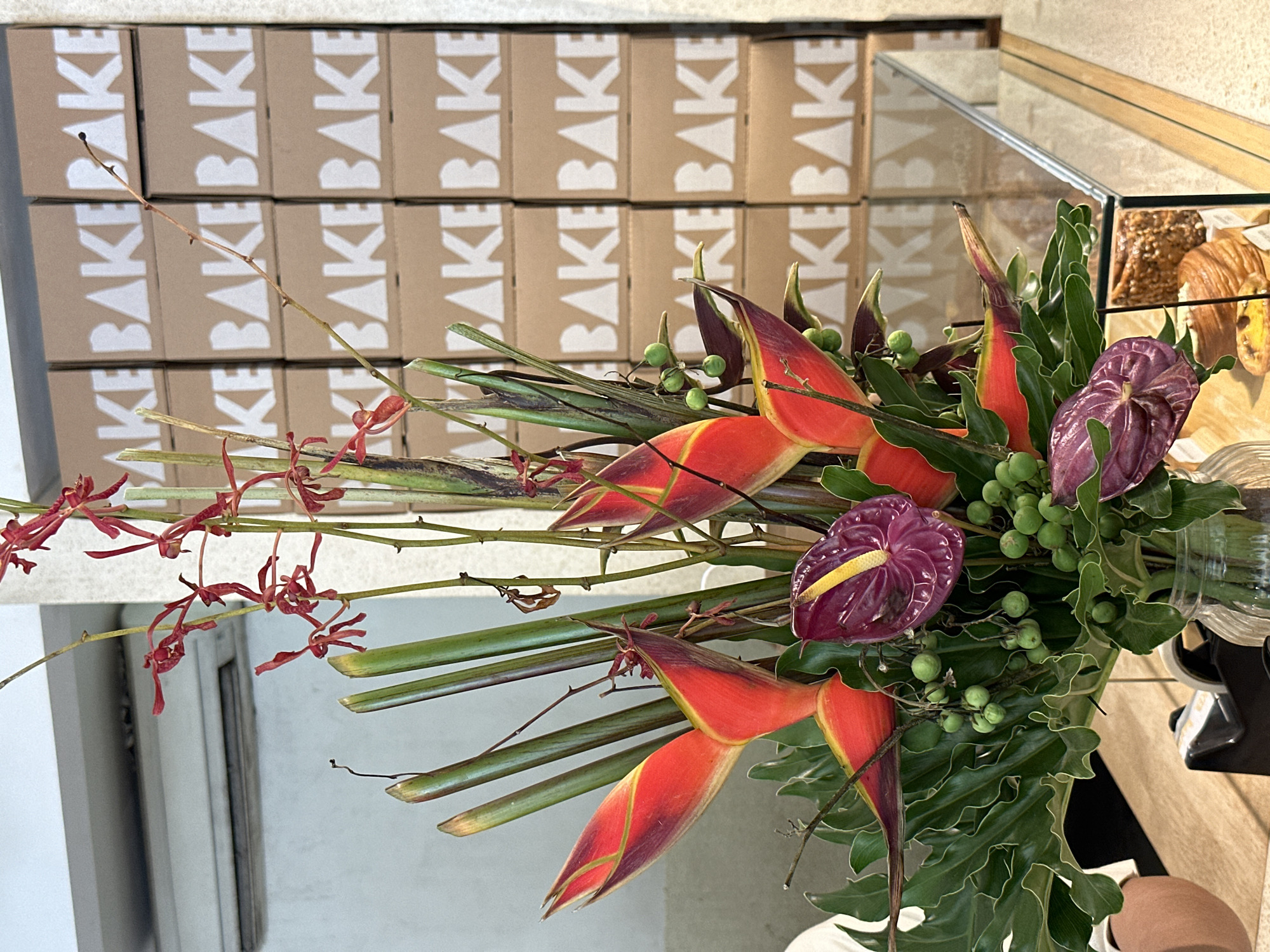 A still life depicts a flower arrangement featuring heliconia and anthurium flowers, set against a backdrop of stacked boxes. The boxes have the word "BAKE" printed on them vertically in large white letters. A glass display case with pastries inside can be seen in the background to the right of the flowers. The scene appears to be located in a bakery or cafe, based on the branding of the boxes and the presence of pastries. The city and specific location are unknown.