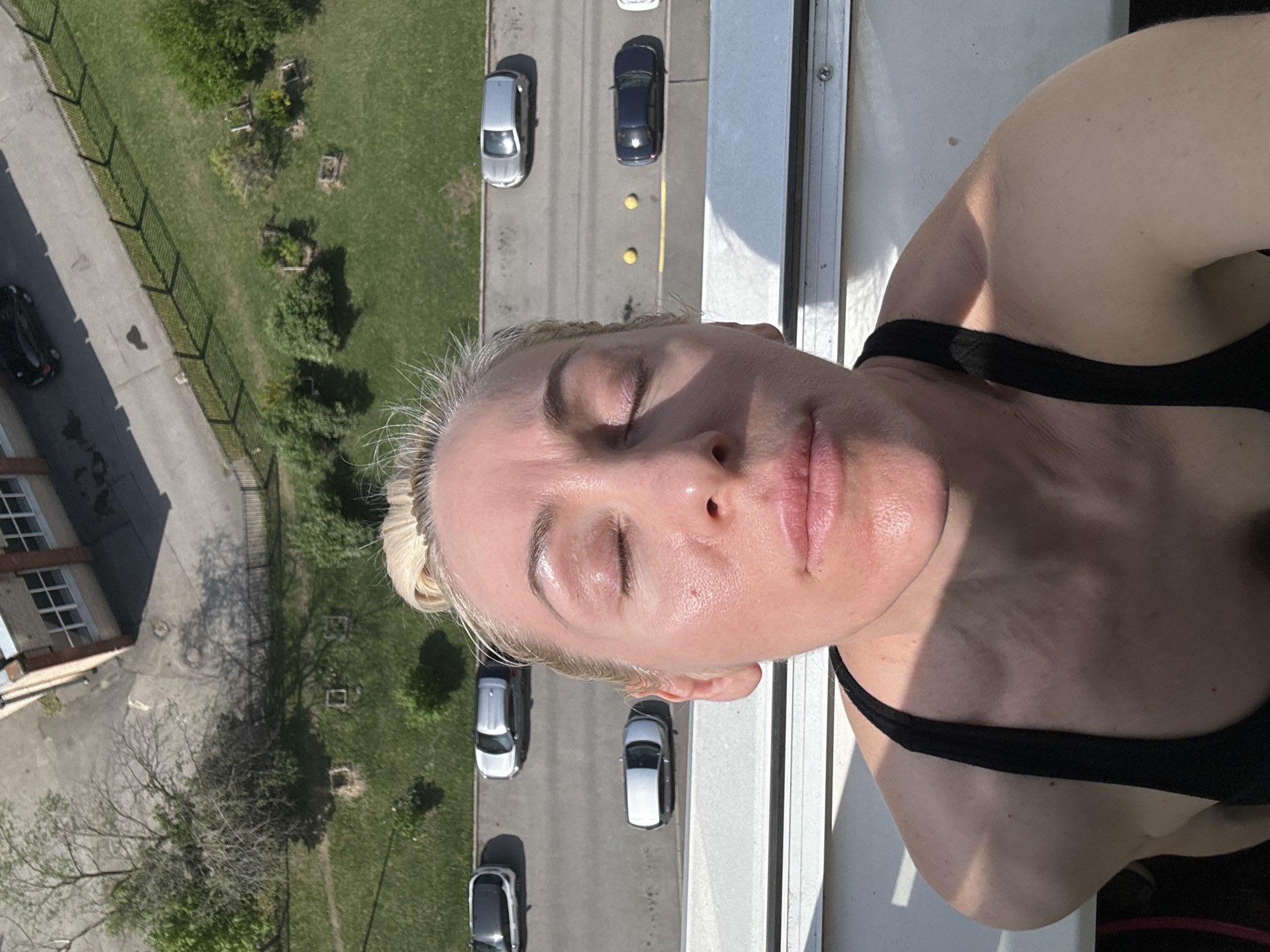 A woman with blonde hair stands on a balcony. She wears a black tank top. The view from the balcony is of a street with parked cars, a building, and a grassy area with trees. There are yellow lane dividers visible on the street.