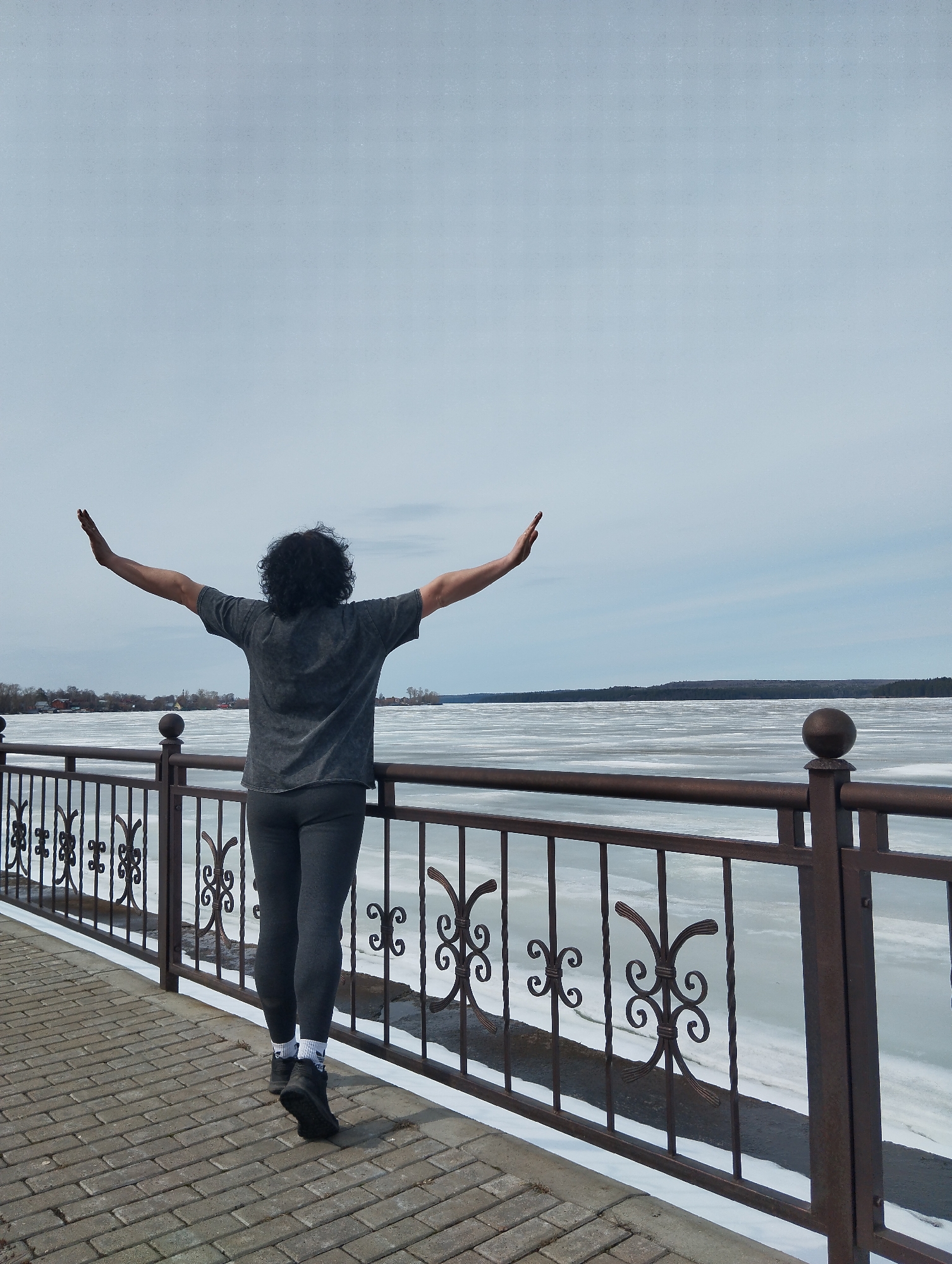 The image depicts a person standing with arms outstretched at a lakeside location, likely in Votkinsk, Russia. The person is facing away from the viewer, wearing a grey t-shirt and leggings, with dark curly hair. They are standing on a paved walkway bordered by an ornate metal railing. Behind the railing, the lake is covered in ice and snow, extending to the horizon. The distant shoreline is visible, with a mix of trees and buildings on the left side. The sky is overcast, suggesting a cold weather.