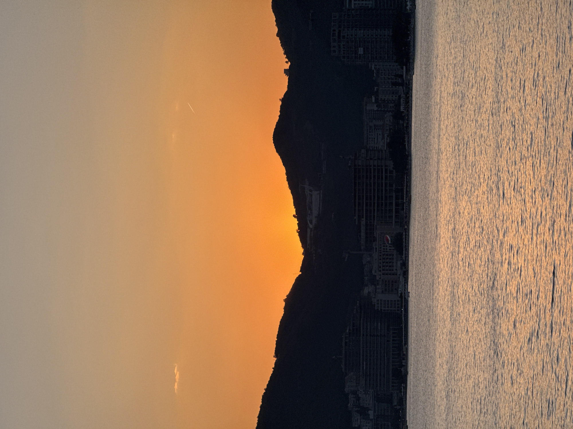 A vertical, wide shot captures a sunset over Budva, Montenegro. The sky is a gradient of soft orange and peach hues, transitioning to a lighter tone towards the top. A small, fluffy cloud drifts in the upper left quadrant, and a faint contrail from an airplane is visible in the upper right.
Silhouetted against the vibrant sky are dark, undulating mountains that form the backdrop of the scene. The setting sun peeks through a gap in the mountains, casting a warm glow. At the base of the mountains, a city skyline emerges. Numerous buildings, appearing modern with multiple floors and dark facades, are visible. Some buildings appear to be under construction, with visible scaffolding. Among the buildings, a flag with red and white markings is discernible.
In the foreground, a large body of water reflects the warm colors of the sunset, creating a shimmering surface with gentle ripples. A few small boats are scattered on the water. The overall impression is one of serene natural beauty juxtaposed with urban development.