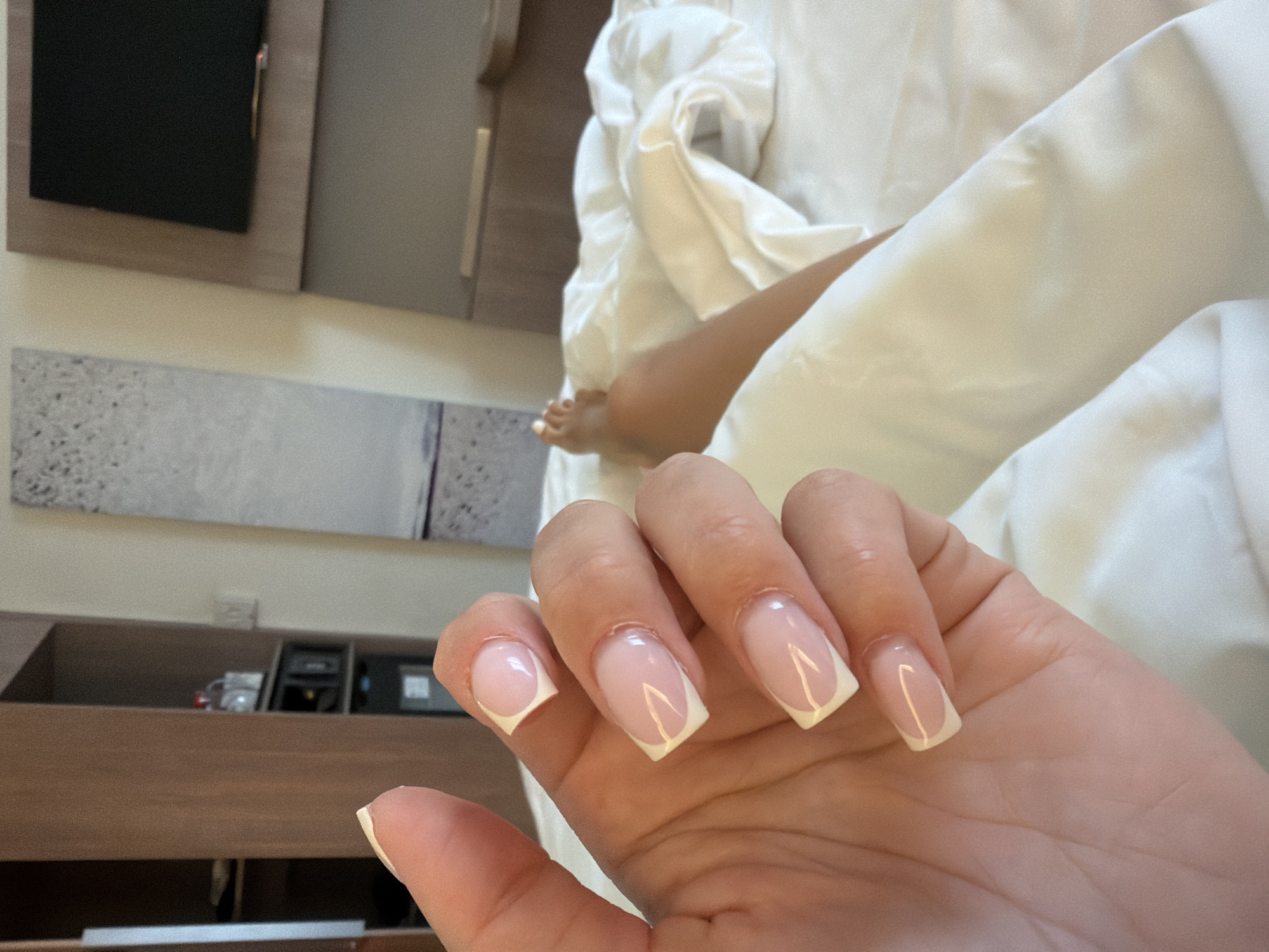 The image presents a close-up view of a person's left hand, showcasing a fresh French manicure. The nails are a square shape with natural pink bases and distinct white tips, some featuring a subtle V-shaped design at the tip.
In the blurred background, part of another person's bare leg and foot are visible, resting amidst rumpled white bedding, indicating a relaxed, indoor setting. Behind the hand, a light brown wooden furniture piece, possibly a dresser or shelving unit, stretches across the frame. Above this, a dark, rectangular screen, likely a television or monitor, is mounted on the wall within a light-colored wooden frame. To the left, a long, narrow piece of abstract art or decorative panel is affixed to the wall. On the shelves of the wooden unit, several small objects, including what appear to be glasses and dark electronic devices, are discernible.
The overall scene suggests a moment of leisure and personal care, likely taking place in a bedroom or hotel room, possibly located in Moscow, Russia. The time of day and weather are not determinable from the indoor lighting. There is no visible text in the image.