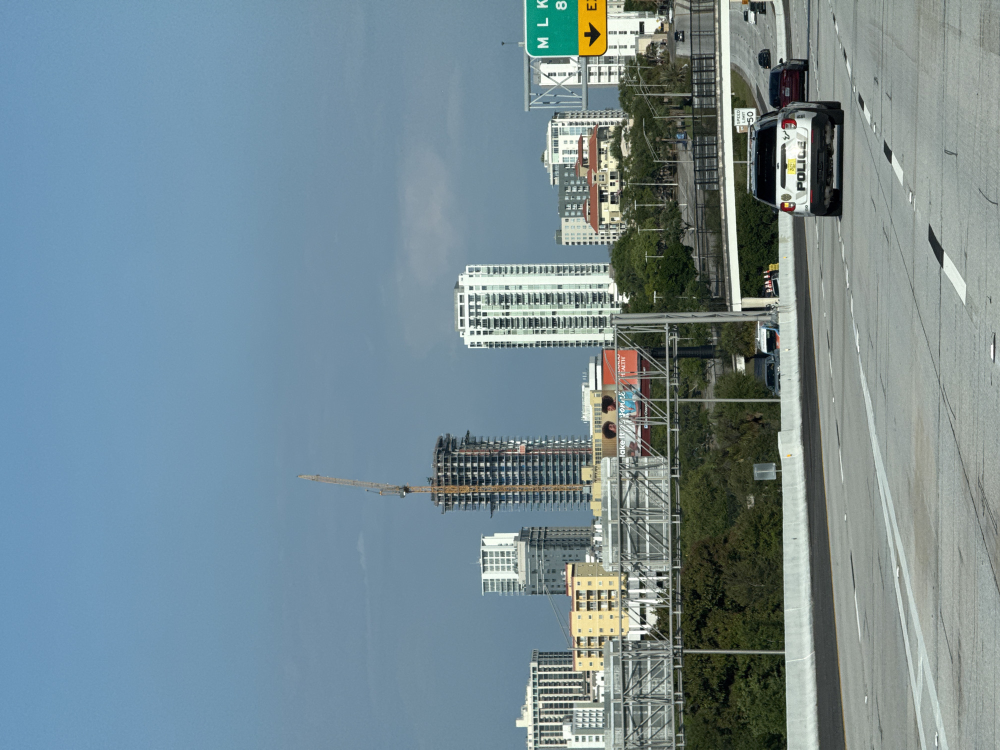 The image depicts a daytime scene on a multi-lane concrete highway, likely I-175 or I-275, looking towards the downtown skyline of Saint Petersburg, Florida, United States of America.
In the foreground, a white SUV police vehicle, viewed from the rear, travels in the right-center lane. The word "POLICE" is visible on its liftgate, and a yellow Florida license plate with partial digits "24X91" is mounted. Other vehicles, including a red sedan and a dark sedan, are present on the highway ahead of the police vehicle. Lane markings consist of solid and dashed white lines. A concrete barrier runs along the right side of the highway, separating it from an area with green foliage.
The midground features an overhead steel truss structure, possibly supporting signage, positioned above the highway. Behind this, lush green trees and an elevated concrete overpass are visible, with a "SPEED LIMIT 50" sign mounted on an overhead gantry. Partial views of commercial billboards are also present behind the truss structure.
The background is dominated by the city skyline under a partly cloudy blue sky. A prominent, unfinished high-rise building with exposed concrete structure and numerous floors is under active construction, featuring a tall yellow tower crane extending vertically alongside it. To its right, several completed white high-rise residential buildings with visible balconies stand. Other mid-rise and lower buildings are scattered throughout the cityscape. On the far right, a green highway exit sign displays "MLK 8TH AVE" and a yellow downward arrow, indicating an exit for Martin Luther King Jr. Street and 8th Avenue.