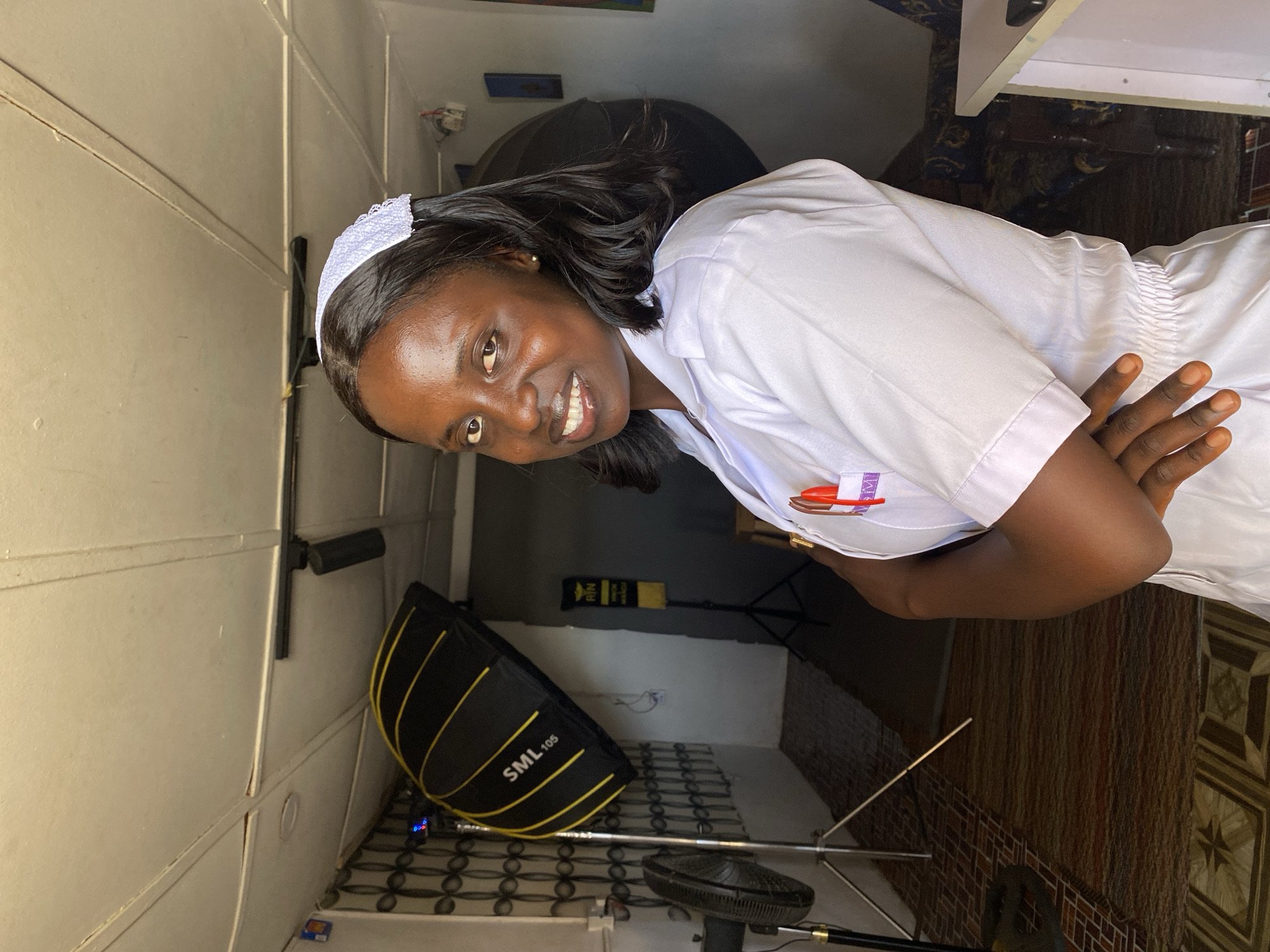 A young woman wearing a white nurse's uniform and cap is photographed indoors. She has dark hair and skin, and she's smiling slightly at the camera. Her arms are crossed. Behind her are a softbox with "SML105" printed on it, a fan, and other photography equipment, indicating a studio or controlled setting. The walls are paneled, and the floor is tiled with a patterned design. The location is identified as Vom, Nigeria.