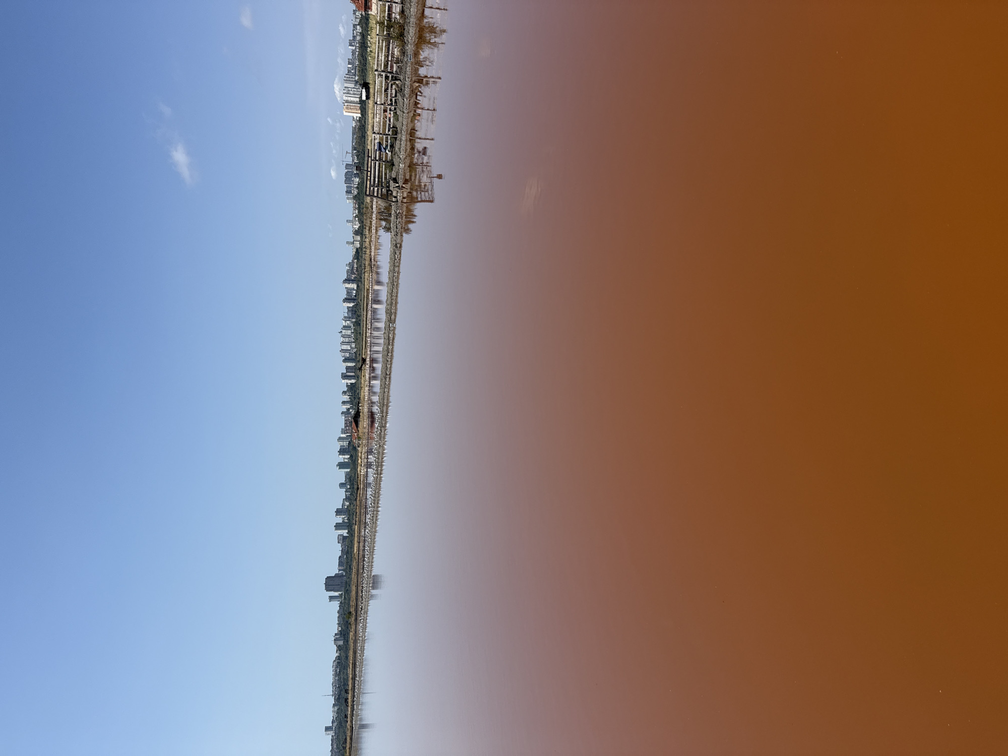 The image depicts the Burgas Salt Pans, a significant natural and industrial site located on the outskirts of the city of Burgas, Bulgaria. The foreground is dominated by a large expanse of reddish-brown water, characteristic of hypersaline environments where Dunaliella salina algae thrive, imparting the distinctive color. In the middle ground, a narrow strip of land separates this water body from another shallow, pinkish water area. This strip is lined with wooden fences and what appear to be salt-drying beds or processing facilities. Further in the background, a dense urban skyline of Burgas is visible, characterized by numerous apartment buildings and skyscrapers, suggesting proximity to the city center. The sky is clear and blue, with a few wispy clouds. The scene suggests a sunny day, with the water's color being the most striking feature. There are no discernible individuals or specific actions taking place in the immediate vicinity of the salt pans, presenting a static, environmental portrait.