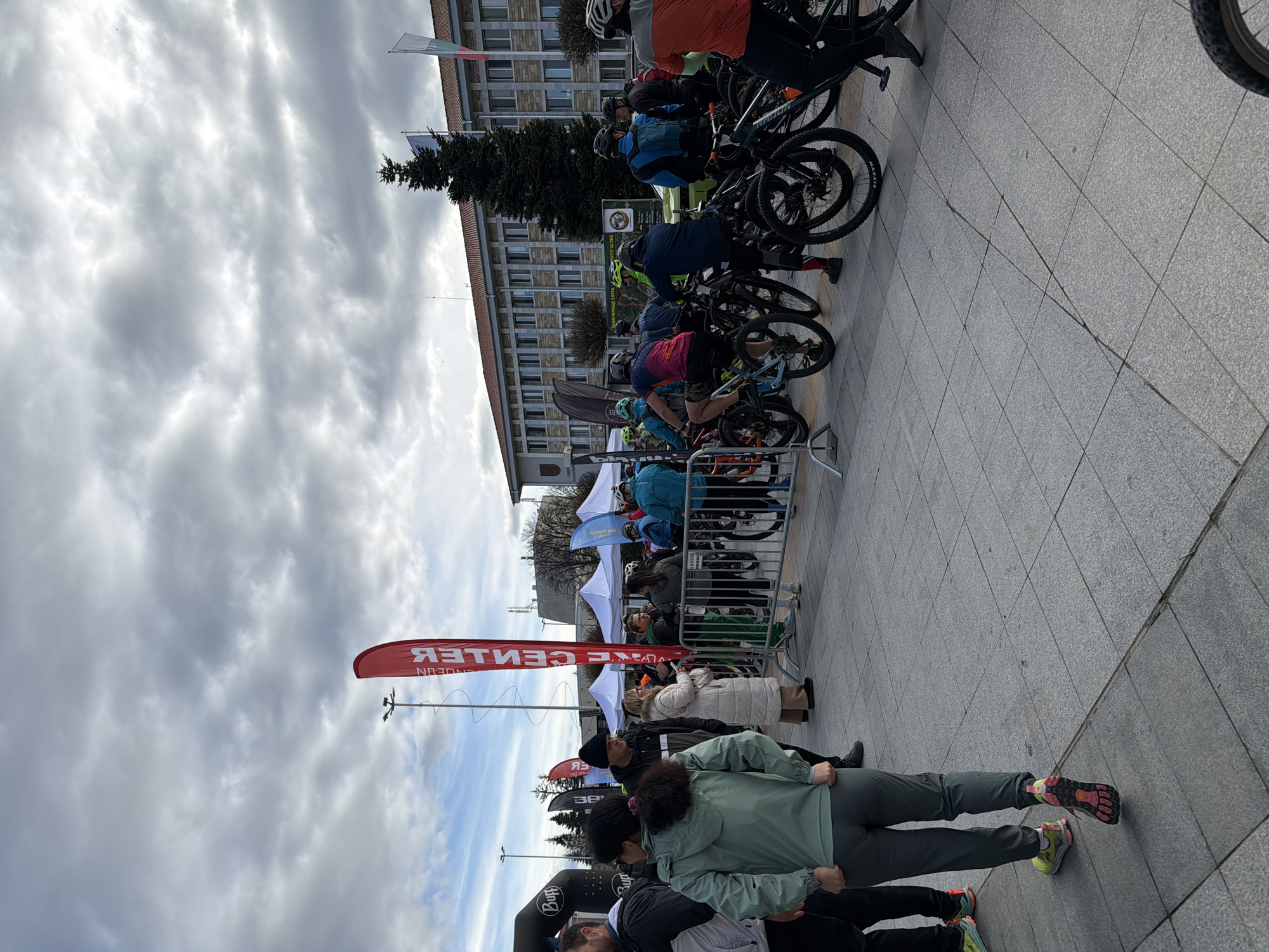 The image shows a gathering of people and bicycles on a paved public square in the city of Panagyurishte, Bulgaria. A group of cyclists is lined up behind a metal barrier, presumably preparing for an event or race, as indicated by the 'CENTER' branded flag. In the foreground, individuals are standing and observing. Behind the cyclists is a building with flags, including the Bulgarian flag. The sky is overcast with thick clouds.