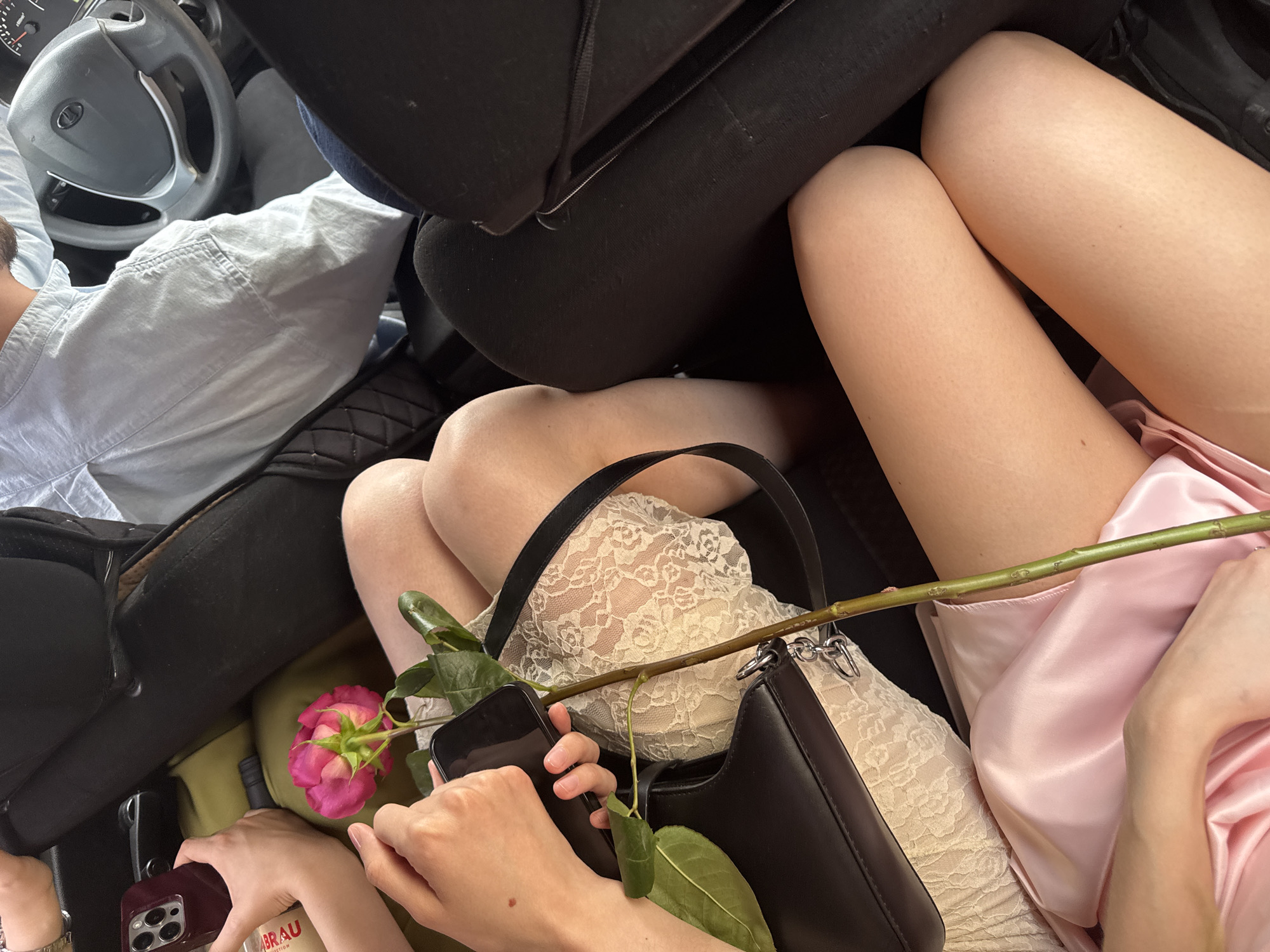 Inside a car, a driver (seen in a light blue shirt and with hands on the steering wheel) is positioned to the left. The focus is on two other people in the back. One individual wears a lace dress, and the other wears a light pink satin garment. A single pink rose rests across their laps, and both individuals hold smartphones. A black purse rests on the lap of the person in the lace dress. Luggage, partially visible in olive green, is present below. The location is indicated as Perm, Russia, by the writing on one of the visible objects in the image (likely a bottle of beer).