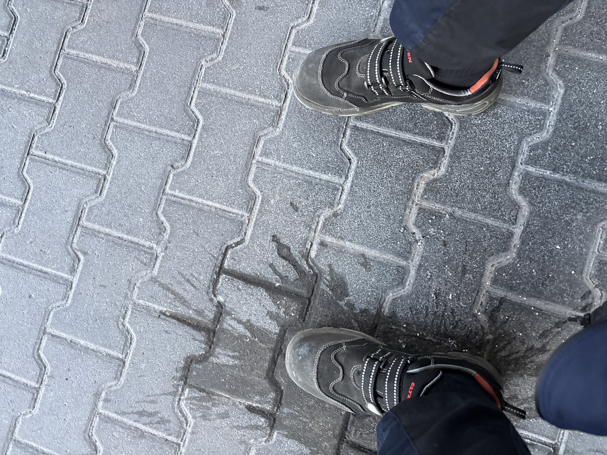 The image shows a pair of black work boots and dark blue pants standing on a wet, interlocking brick surface in Mannheim, Germany. The boots have white stitching and strap closures. The ground appears to be an outdoor walkway, possibly a street or sidewalk, with visible water pooling and reflecting light on the bricks. The overall lighting suggests it is daytime.