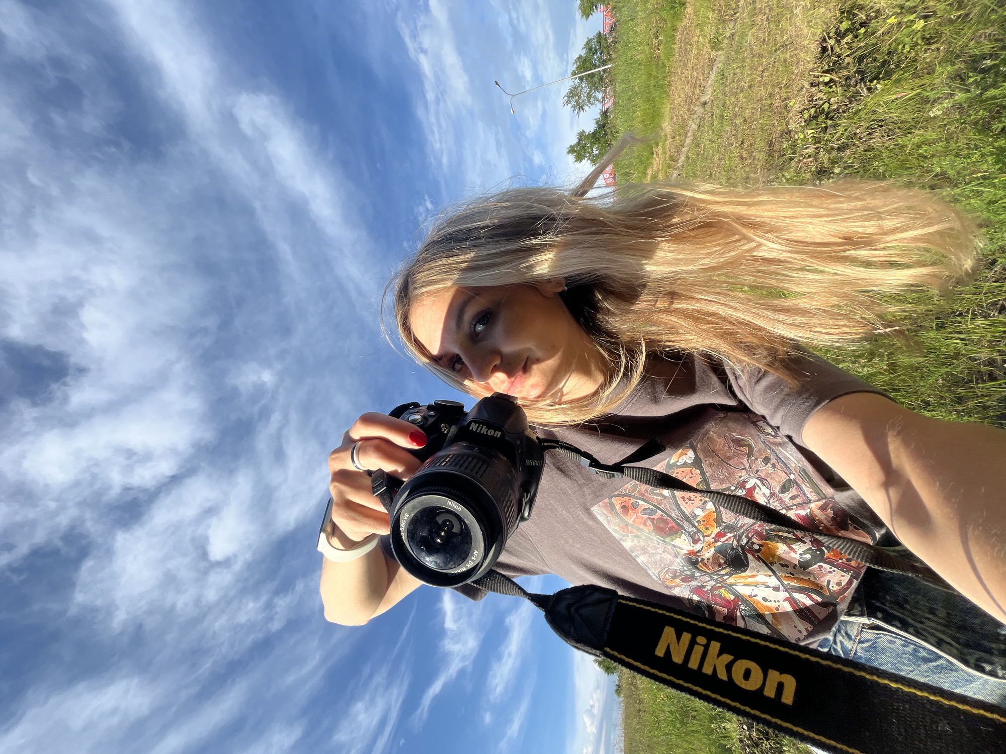 The image shows a young woman holding a Nikon camera and taking a selfie outdoors. She has long blonde hair and is wearing a grey t-shirt with a colorful graphic print and blue jeans. The camera strap, with the Nikon logo visible, is around her neck. She is wearing a ring on her left hand and a smartwatch on her left wrist. The background is a grassy field under a bright blue sky with wispy clouds. In the distance, there are trees, a red bridge, and a lamppost. The scene appears to be a rural or park area. The lighting suggests it is daytime, likely late afternoon due to the warm tones and shadows. The overall atmosphere is casual and active, with the woman seemingly engaged in photography. The location is identified as being in the city of Chaikovsky, Russia.
