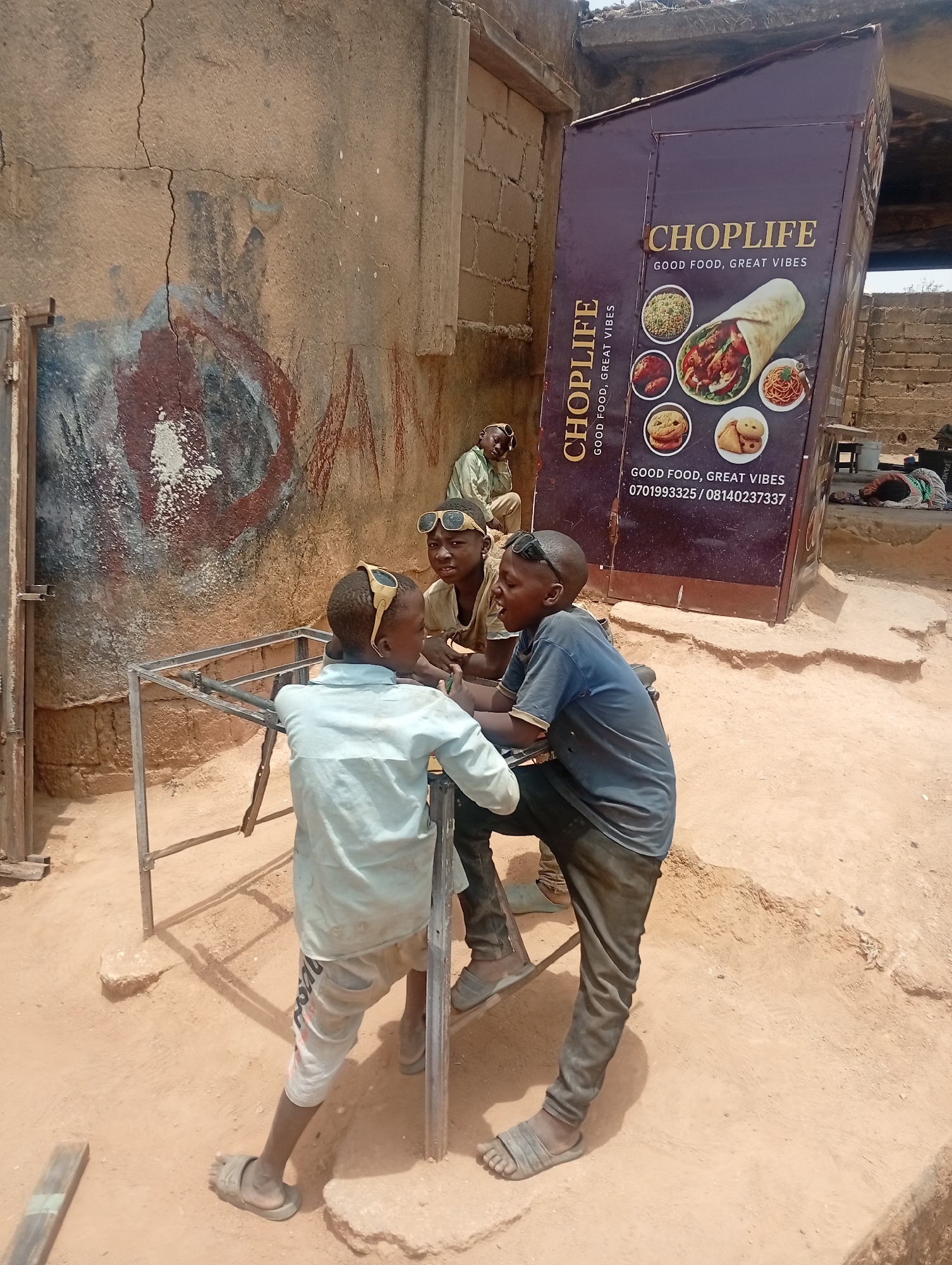 An outdoor scene in Jos, Nigeria, features three young boys gathered around a metal frame, possibly an unfinished table, under bright daylight. The boys, wearing casual shirts, trousers or shorts, and sandals, appear to be conversing or interacting. Two of them have sunglasses pushed up on their heads. Their expressions suggest engagement and casualness, with one boy in a blue shirt appearing to speak animatedly. In the background, another person is seen resting or sleeping on the ground near a makeshift structure.
The setting is characterized by unpaved, reddish-brown, dusty ground and an old, cracked wall behind the boys, which bears faded, abstract reddish and bluish markings. To the right, a prominent purple food stand or kiosk is angled, displaying the brand "CHOPLIFE" with the slogan "GOOD FOOD, GREAT VIBES." The stand features images of various food items, including a large wrap, rice, chicken, cookies, noodles, and samosas. Visible text on the stand includes "CHOPLIFE," "GOOD FOOD, GREAT VIBES," and two phone numbers: "0701993325" and "08140237337." The harsh shadows indicate a sunny, dry day, possibly around midday. The overall atmosphere suggests a casual, possibly informal commercial or social gathering area.