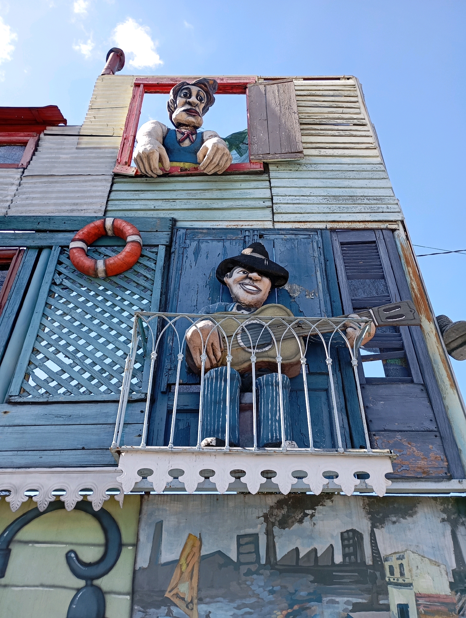 The image depicts the exterior of a building in La Boca, Buenos Aires, Argentina, characterized by its vibrant and eclectic architectural style. The building features multiple levels with distinct facades constructed from various materials, including corrugated metal, weathered wood planks painted in shades of blue, red, and off-white, and metal sheeting.
Dominating the upper portion of the image is a wooden sculpture of a man peering out of a red-trimmed window. He has a gaunt face with wide eyes and is wearing a blue vest and a red scarf. His large, rough-textured hands rest on the window ledge. Above and to the left of this sculpture, a red-topped chimney extends upwards against a clear blue sky with sparse white clouds.
Below this, on a lower level, a second sculpture of a man is positioned on a balcony. This figure wears a black hat and a blue shirt with vertical stripes, and he is depicted holding and playing an acoustic guitar. The balcony is adorned with decorative white metal railings and trim. Behind the figure, the wall is made of weathered blue wooden doors and shutters. To the left of the balcony, a bright orange life preserver with white stripes is affixed to the wall, overlaying a blue lattice structure.
The lowest visible section of the building displays a mural. This mural appears to depict an industrial or port scene with smokestacks, buildings, and a sailboat. The overall impression of the building is one of artistic and whimsical construction, typical of the colorful Caminito street in La Boca. The lighting suggests it is daytime, with bright sunlight casting shadows and illuminating the scene.