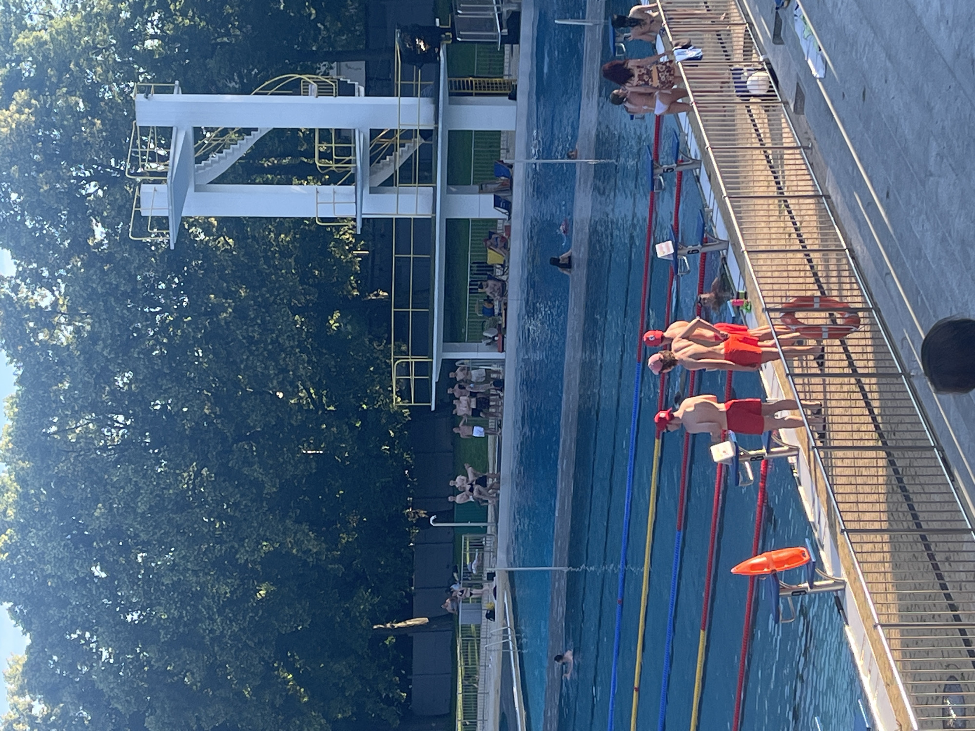 The image shows an outdoor swimming pool with a diving platform. Several people are present around and in the pool. In the foreground, four young men wearing red swim trunks and caps stand on the pool deck. One of them is holding a white object. An orange rescue buoy is attached to the railing in front of them. In the middle ground, there are several lanes marked in the pool with buoys, and some people are swimming. On the far side of the pool, near the diving platform, a group of people are gathered. The diving platform is tall and white with yellow railings, and a spiral staircase leads up to it. The background consists of a dense line of trees. The lighting suggests it is a sunny day, with shadows cast on the pool deck. The overall scene depicts a public swimming pool facility.