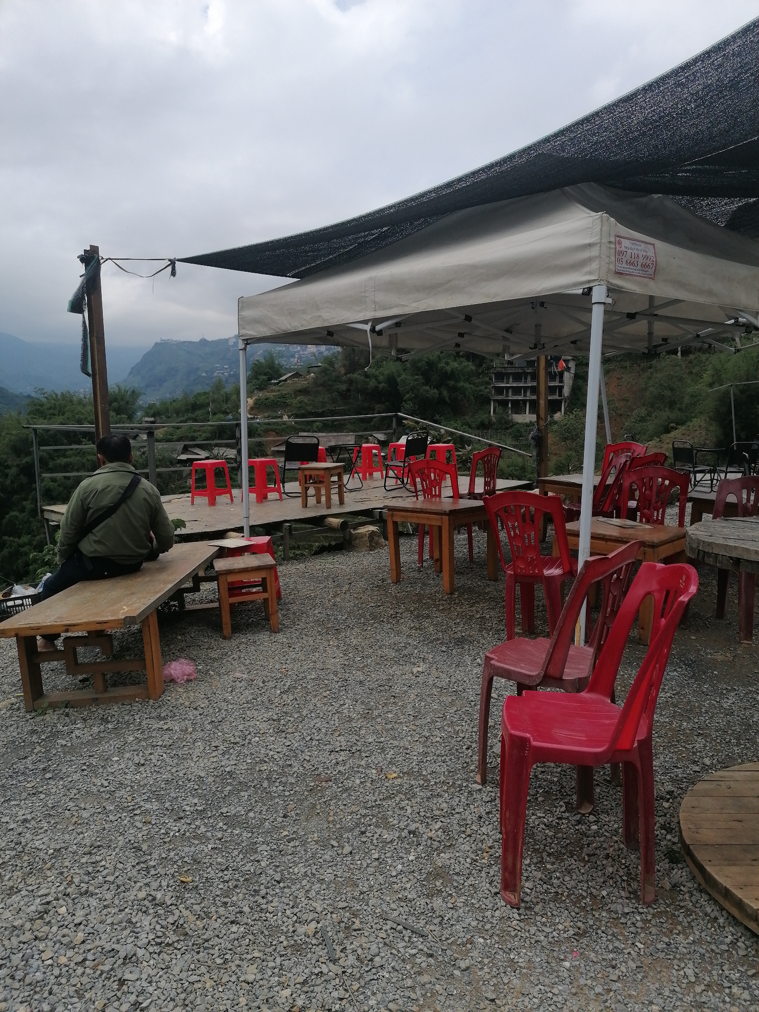 The image depicts an outdoor seating area in Cat Cat Village, Vietnam. A man in a green jacket sits on a wooden bench, facing away from the camera, overlooking a mountainous landscape. Tables and red plastic chairs are scattered on a gravel surface under a white tent and a black mesh awning. In the background, buildings are visible on a hillside under a cloudy sky.