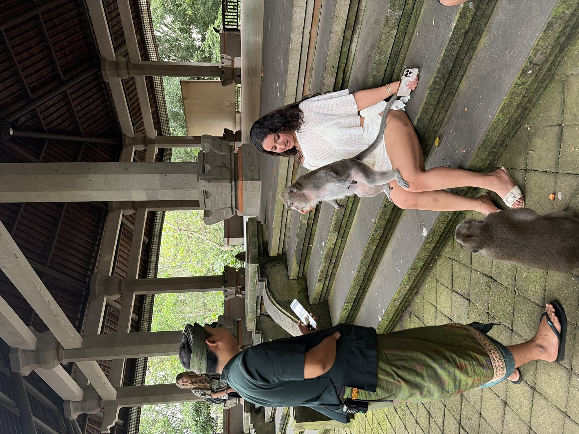 In a courtyard area of a temple in Ubud District, Indonesia, a woman in a white outfit sits on stone steps with a monkey perched on her lap. She is smiling and looking down at the monkey. Another monkey sits at her feet. To the left, a man in a dark green uniform and a patterned sarong is holding a phone and looking at it. Further back and to the left, a woman with long blond hair is standing. The scene is framed by stone pillars and a wooden roof overhead. The steps and ground are covered in moss and some fallen leaves.