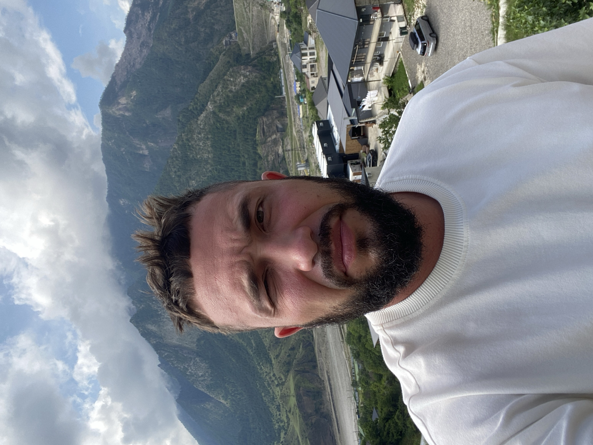 A man with a beard and short, spiky hair is taking a selfie outdoors. He is wearing a white crewneck sweatshirt. Behind him, a mountain range is visible, with some trees and buildings on the slopes. The sky is cloudy. The location is identified as Chalda, Russia.