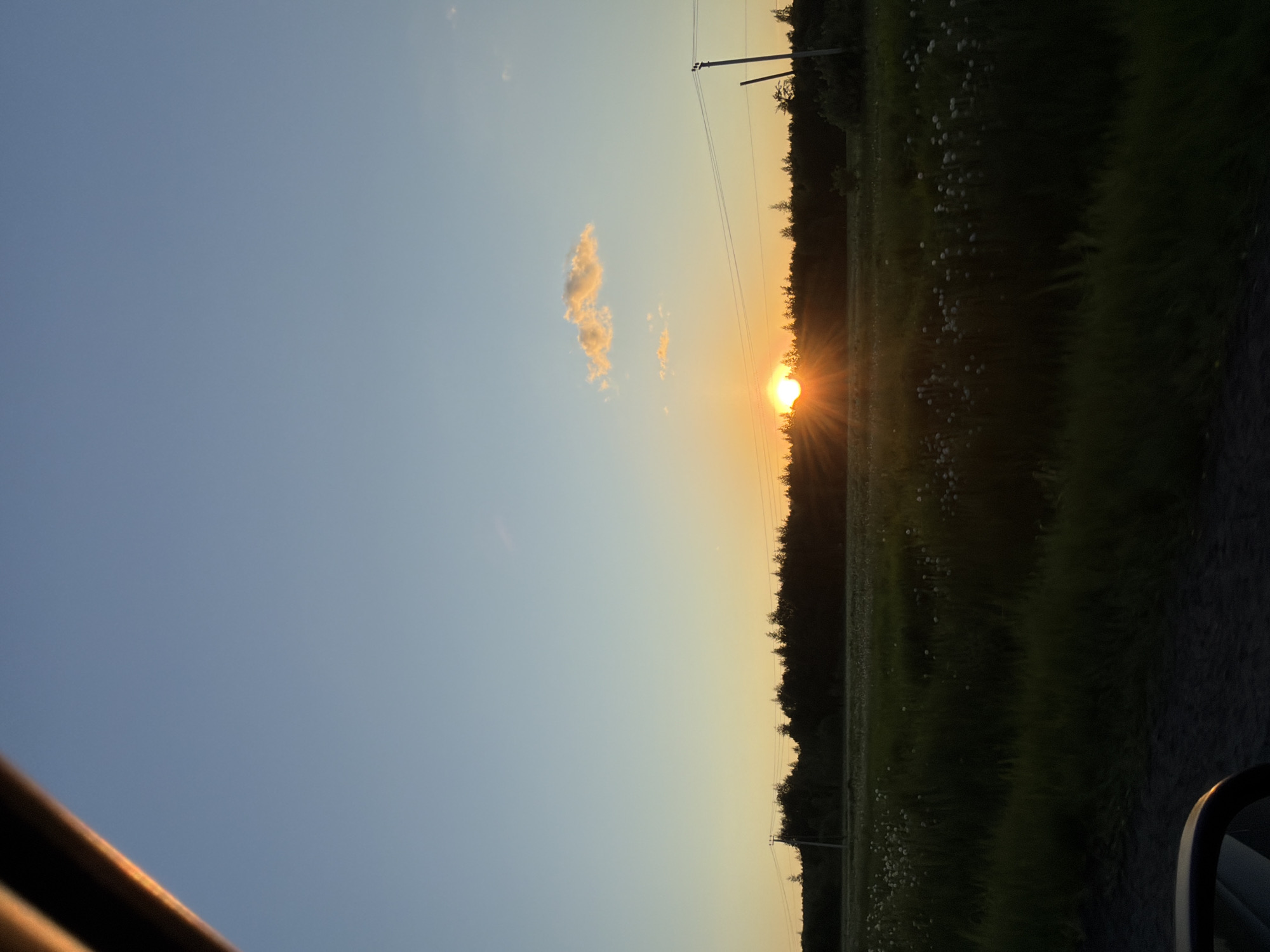 The image depicts a sunset in Mikhalevo, Russia. The sun is setting over a treeline, casting rays of light. A single cloud is visible in the blue sky. Power lines run parallel to the treeline. A grassy field with white flowers stretches from the treeline to the right edge of the image. The edge of a car is visible in the bottom left and bottom right corners.