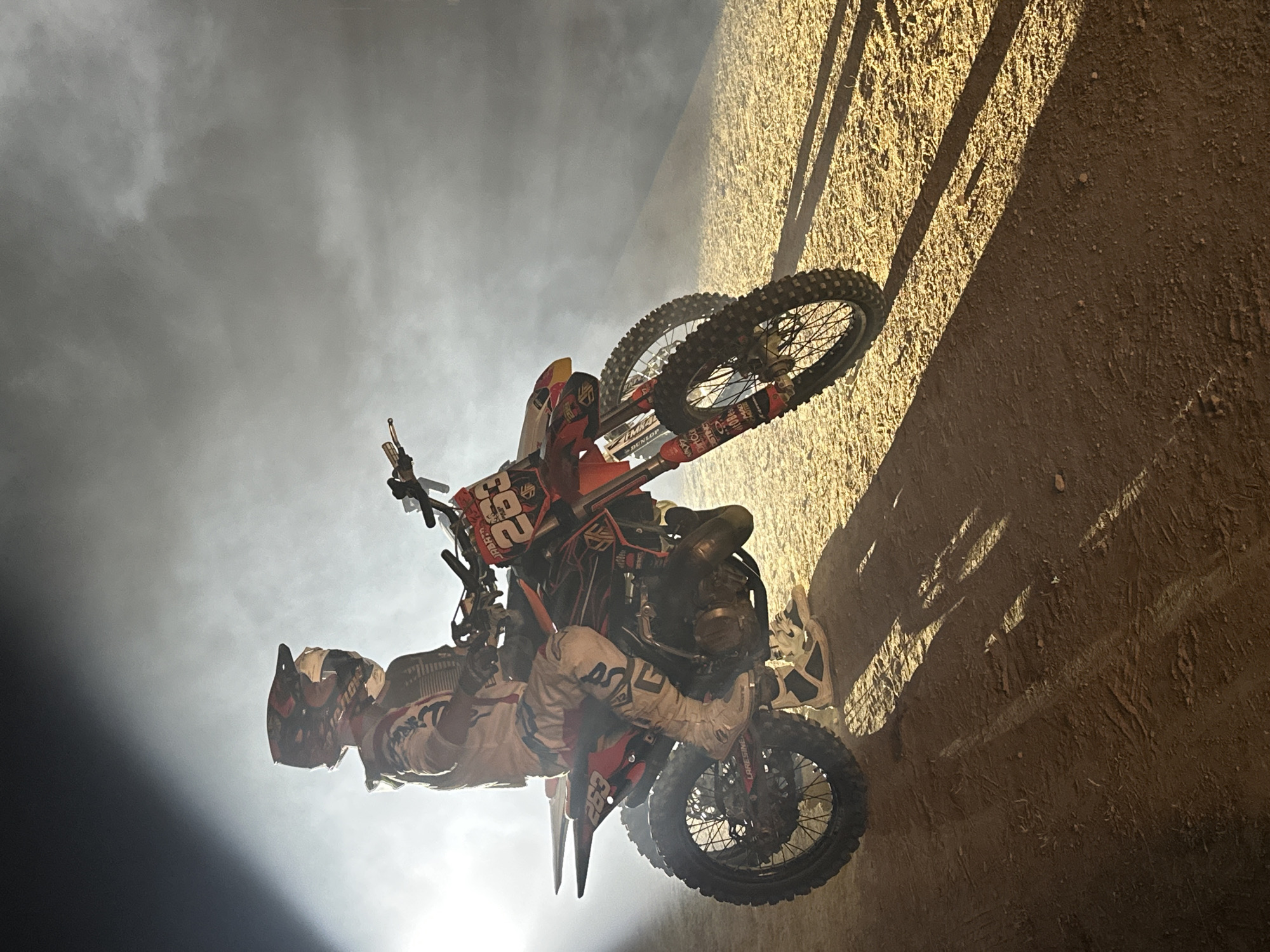 The image depicts a motocross rider posed on a dirt track, illuminated by a bright spotlight from the left, creating dramatic shadows. The rider, wearing full gear including a helmet and racing suit with the number "263" visible on the bike, sits astride a red and black dirt bike. The ground is covered in dirt and small pebbles. Behind the rider, a cloud of smoke or fog is illuminated by the spotlight, obscuring the background. There is no indication of San José del Valle, Spain, within the image itself.