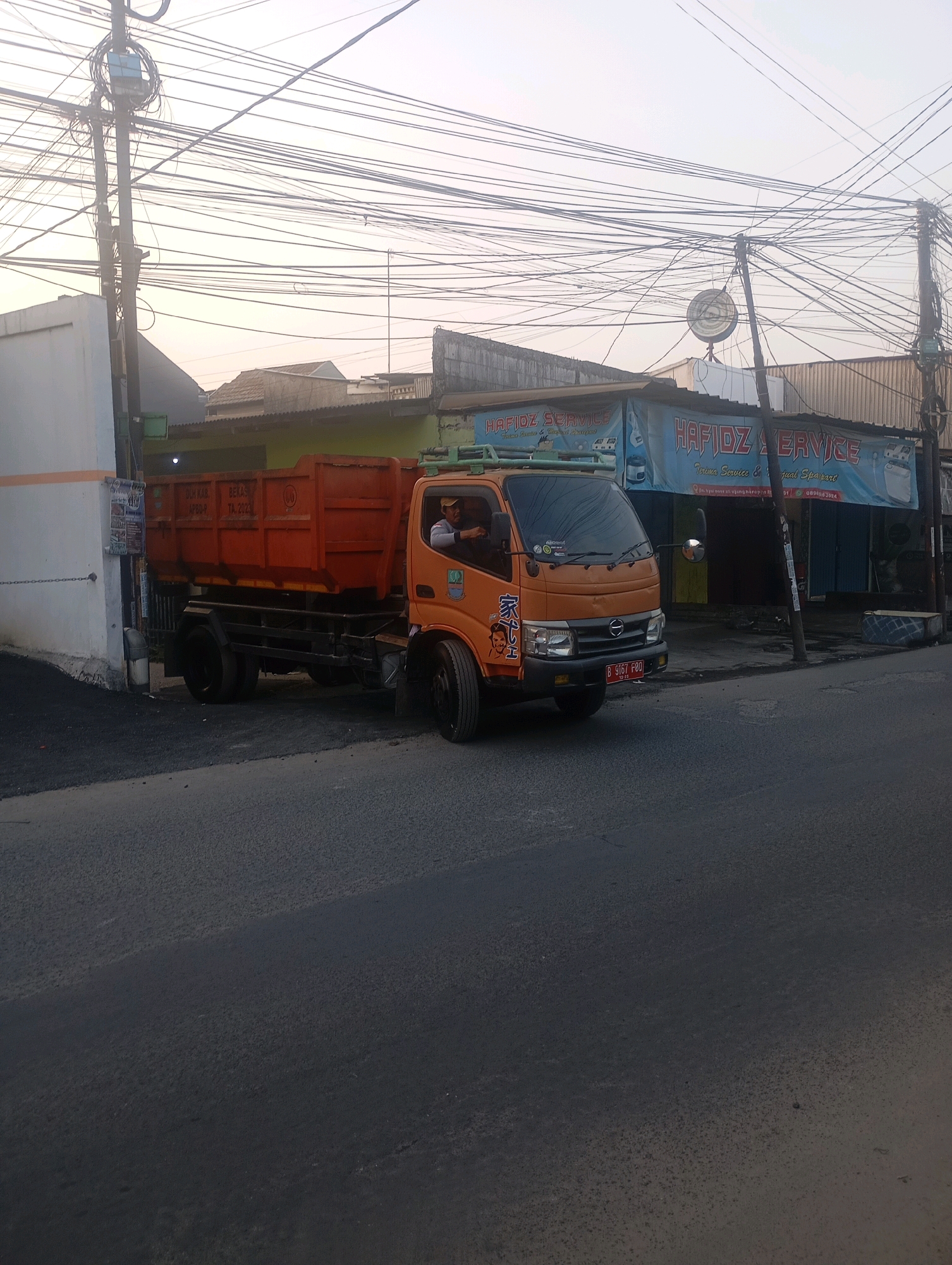 A dump truck is parked on the side of a road in Babelan, Indonesia. The truck is orange and appears to be empty. A man is sitting in the driver's seat. There are several buildings in the background, including a shop with a blue awning that says "HAFIDZ SERVICE". The sky is overcast. The road is paved and there are power lines overhead.