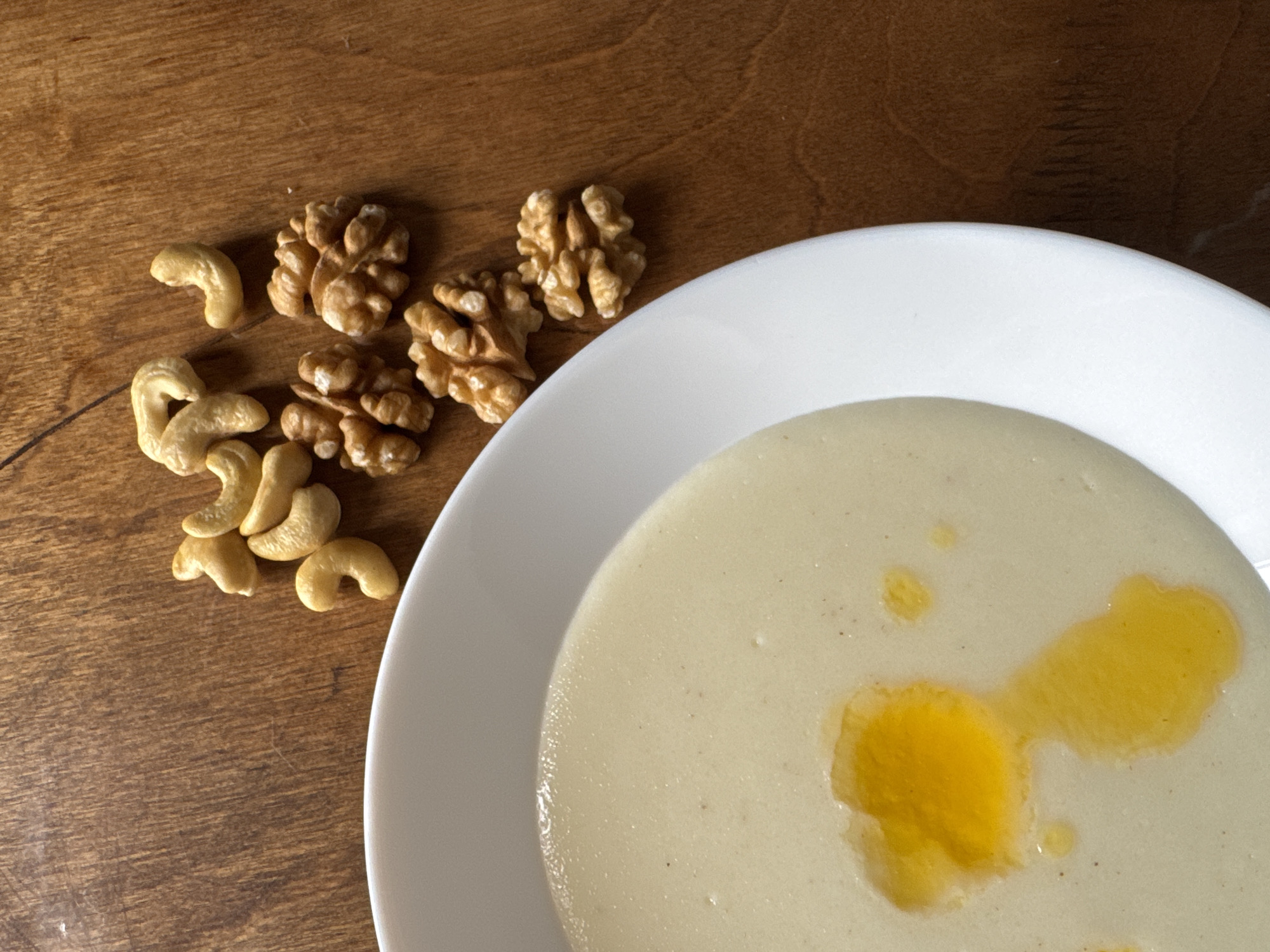 On a wooden surface, a white plate holds a pale-beige cream soup with streaks of yellow oil on the surface. To the left of the plate are several cashew nuts and walnut halves. The environment appears to be a simple, indoor setting. There is no indication of the location being Гаревая, Russia.