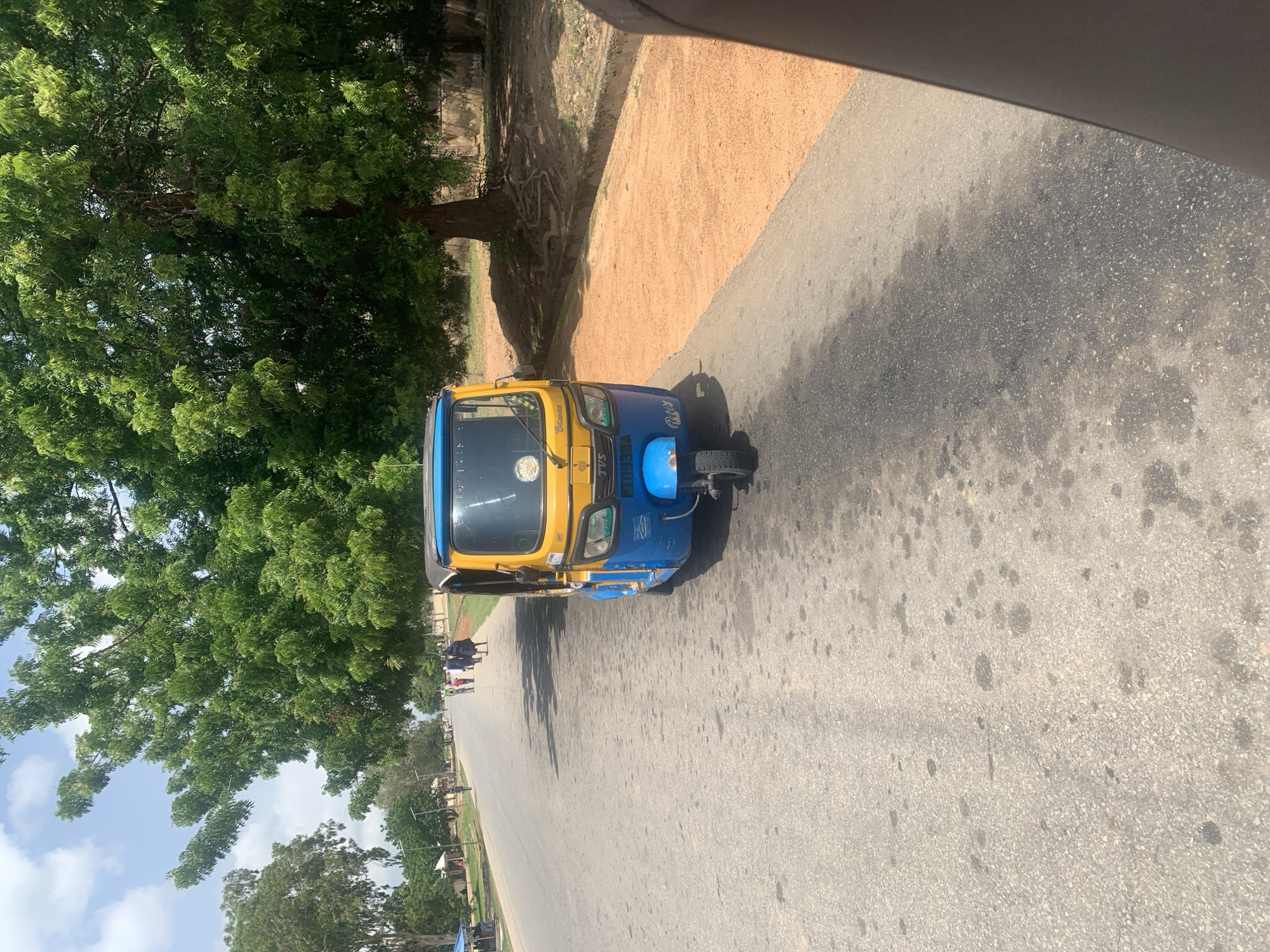 In Jalingo, Nigeria, a bright yellow and blue auto-rickshaw is parked on the right side of a paved road. The auto-rickshaw, a common mode of transport in the region, has a blue front fender and a yellow body with blue accents. Its windshield is visible, and a sticker is affixed to the right side of the dashboard. The road surface is grey asphalt with scattered dark stains, possibly oil or water marks, and appears well-trafficked, with tire tracks visible.
To the left of the road, a line of trees with lush green foliage creates a shaded canopy, suggesting a warm climate. A few individuals, dressed in casual attire, are walking along the left side of the road in the distance, their figures appearing small against the vastness of the landscape. Some buildings or structures are partially visible behind the trees.
The right side of the road features a strip of reddish-orange soil, contrasting with the grey asphalt. A large tree with exposed roots stands prominently on this side, its branches extending over the area. The sky above is a pale blue with scattered white clouds, indicating daytime and fair weather. A portion of a dark, blurry object, possibly the interior of a vehicle or a person's clothing, occupies the extreme right edge of the frame, suggesting the photo was taken from a moving vehicle or from within another object. The overall scene depicts a typical roadside environment in Jalingo, characterized by a mix of urban transportation, natural elements, and pedestrian activity.