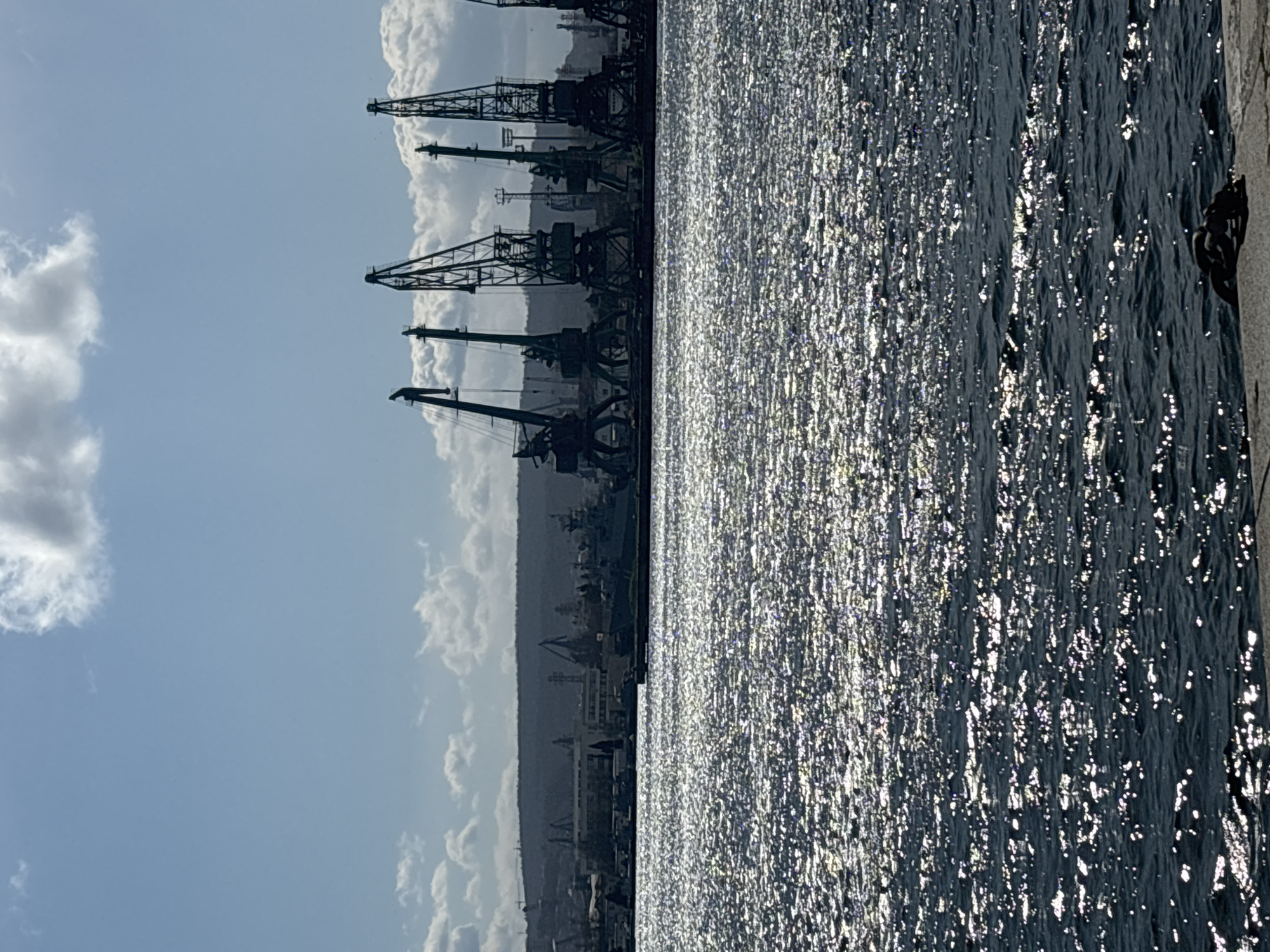 This photograph captures a scene in Varna, Bulgaria, featuring a body of water, likely the Black Sea, reflecting sunlight intensely, and industrial cranes on the horizon. The sky above displays a clear blue with scattered white clouds. The foreground is dominated by the shimmering water, while the skyline is marked by numerous cranes, suggesting a port or industrial area. There are no visible people or immediate actions occurring in the shot. The environment is a mix of natural (sky, water) and man-made (cranes, harbor structure) elements, indicative of an urban coastal setting.