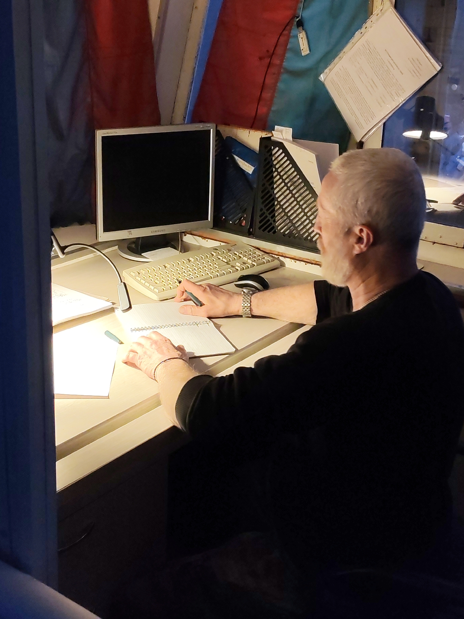 A man with white hair is seated at a desk, writing in a notebook with a pen in his right hand. He is wearing a black long-sleeved shirt and a watch on his left wrist. A computer monitor, keyboard, and desk lamp are on the desk. Behind the desk are file organizers and a document taped to the wall. The location cannot be determined from the image.