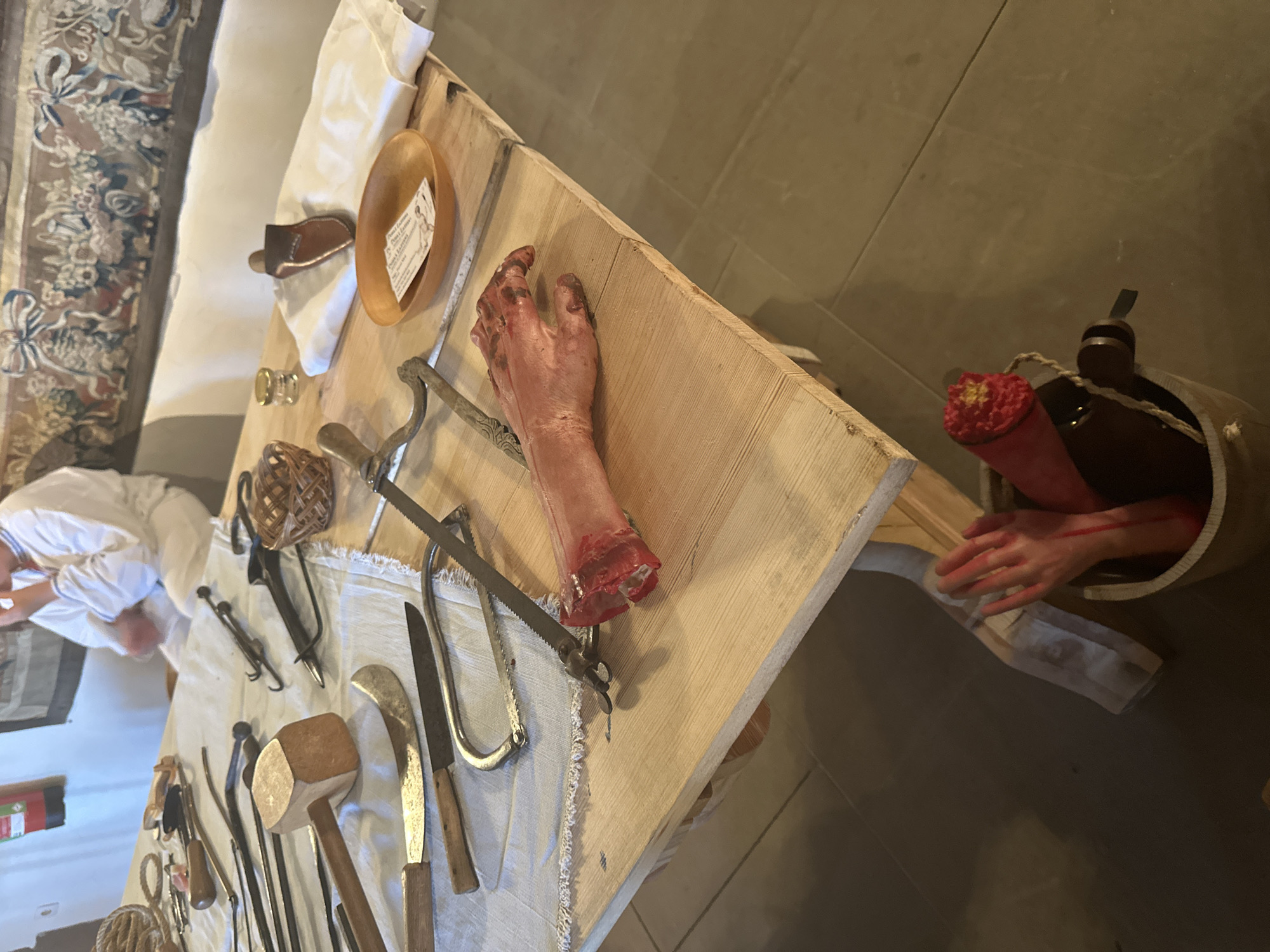 The image depicts a table laid out with various historical medical instruments, including saws, knives, forceps, and a mallet, all placed on a white cloth. In the foreground, a realistic prop of a dismembered arm lies on the table, with bloodied details. Another bucket in the lower portion of the frame also contains dismembered limb props. A person in historical attire is partially visible in the background, suggesting a historical reenactment or museum exhibit. The overall setting appears to be indoors, possibly within the castle of Vianden, Luxembourg, given the tapestry visible in the background.