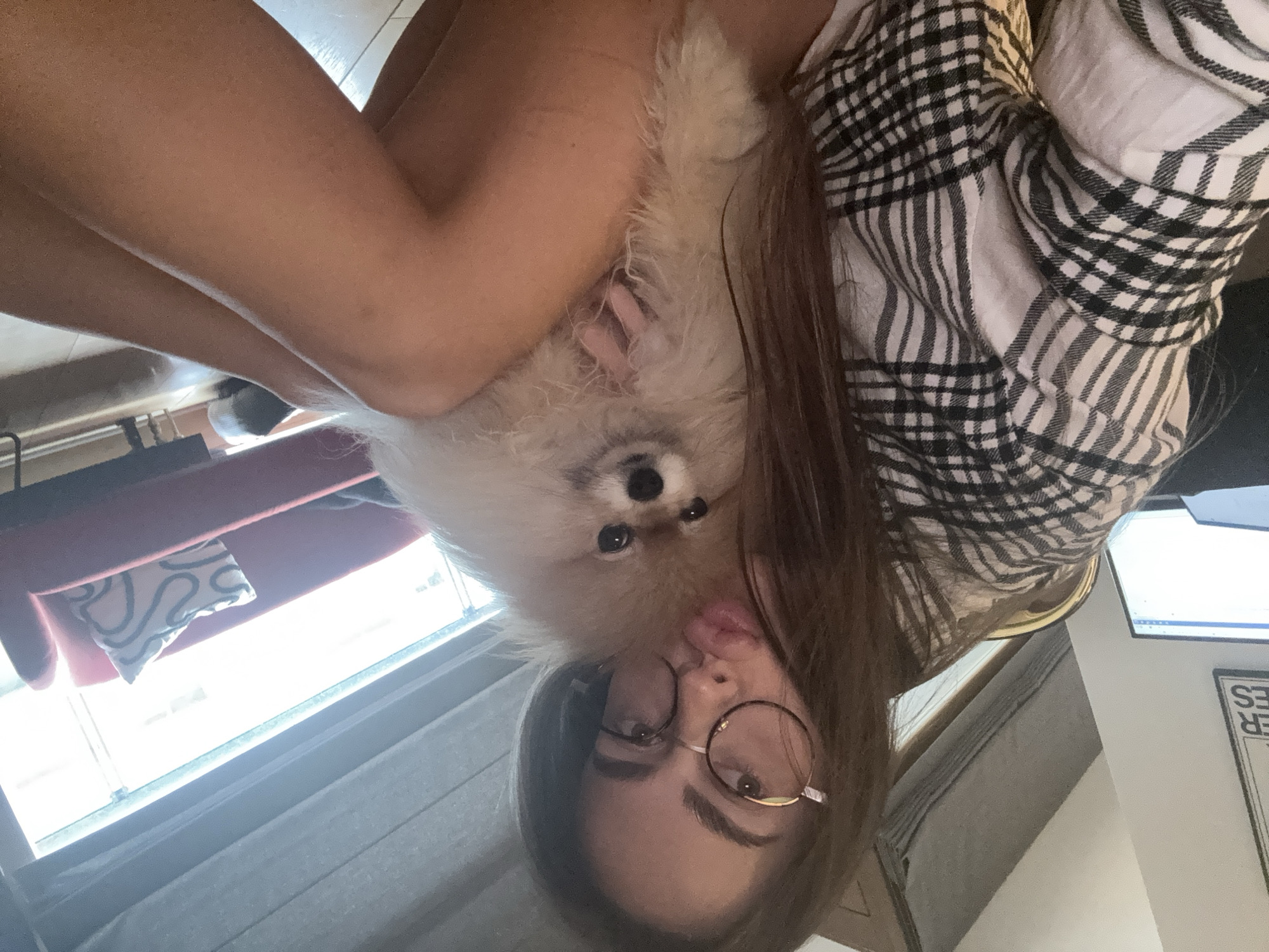 A woman with long brown hair and round eyeglasses is holding a small, fluffy, cream-colored dog close to her face. The dog has dark eyes and a small nose. The woman is wearing a black and white checkered shirt. They are indoors, near a window with blinds and a red cushion. A desk with a computer monitor is visible in the background. It's impossible to determine the precise location within Warsaw, Poland, without further information.