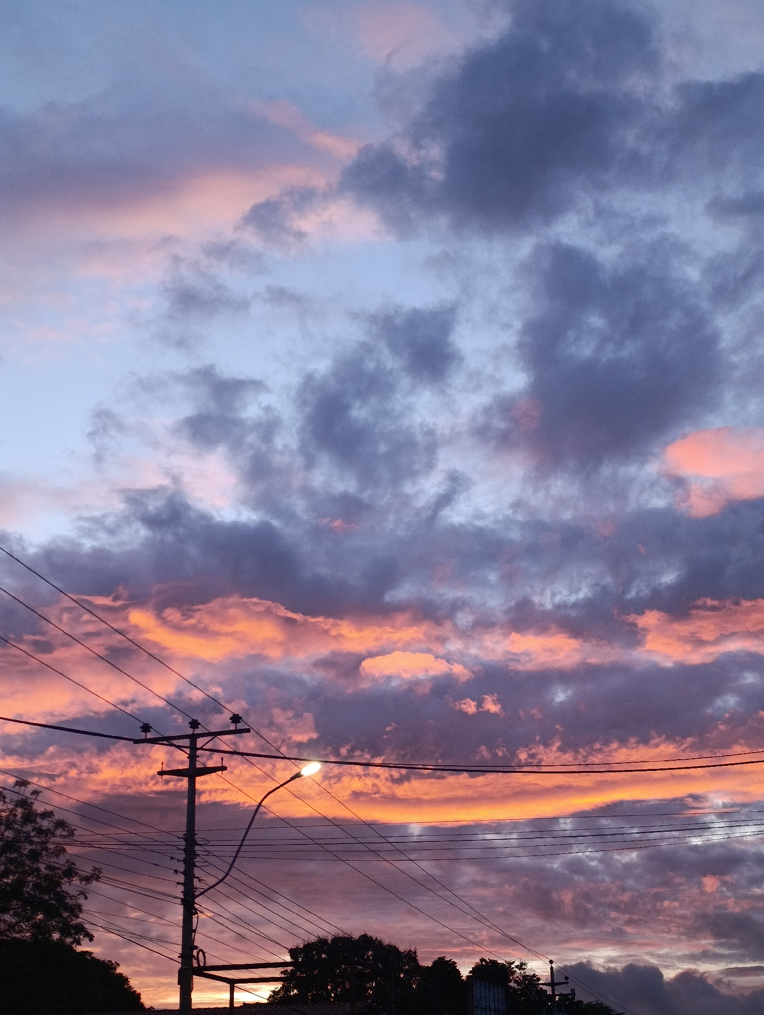 A vertical, eye-level shot shows a dramatic sunset sky above Juan Griego, Venezuela. The sky is a mix of light blue and grey-blue in the upper portion, transitioning to vibrant orange and pink hues near the horizon. Clouds are layered throughout, with darker grey clouds interspersed amongst the brighter sunset colors. In the foreground, a utility pole stands tall, with numerous power lines stretching across the frame. A street light attached to the pole is illuminated. Silhouetted trees and other vegetation are visible at the bottom of the image.