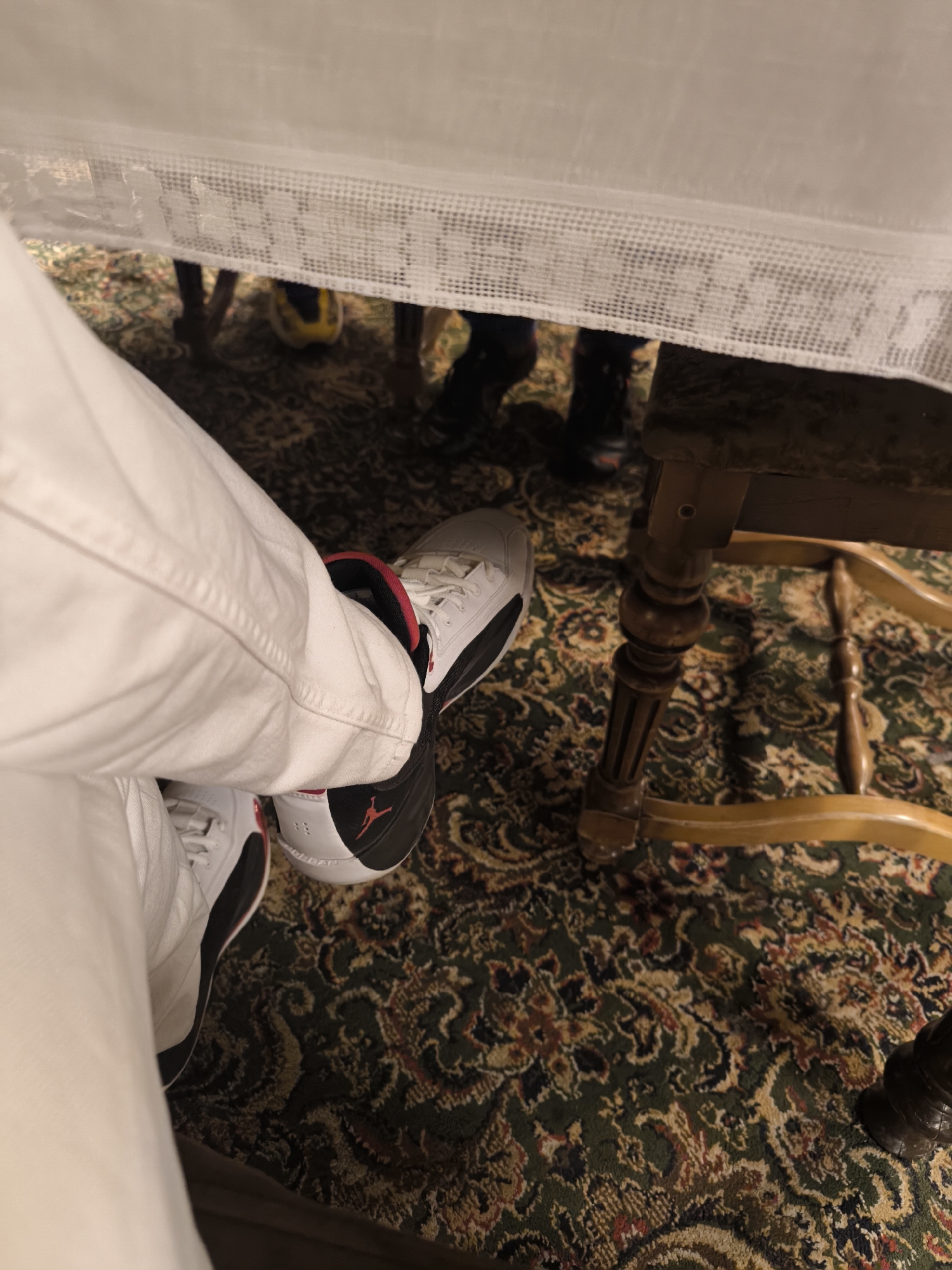 A first-person perspective shot shows a person wearing white pants and black and white sneakers under a table. The shoes have red accents. The table is covered with a white lace tablecloth. The floor is covered with a dark green carpet with an ornate pattern. A wooden chair leg is visible to the right. In the background, the feet of other people are visible.